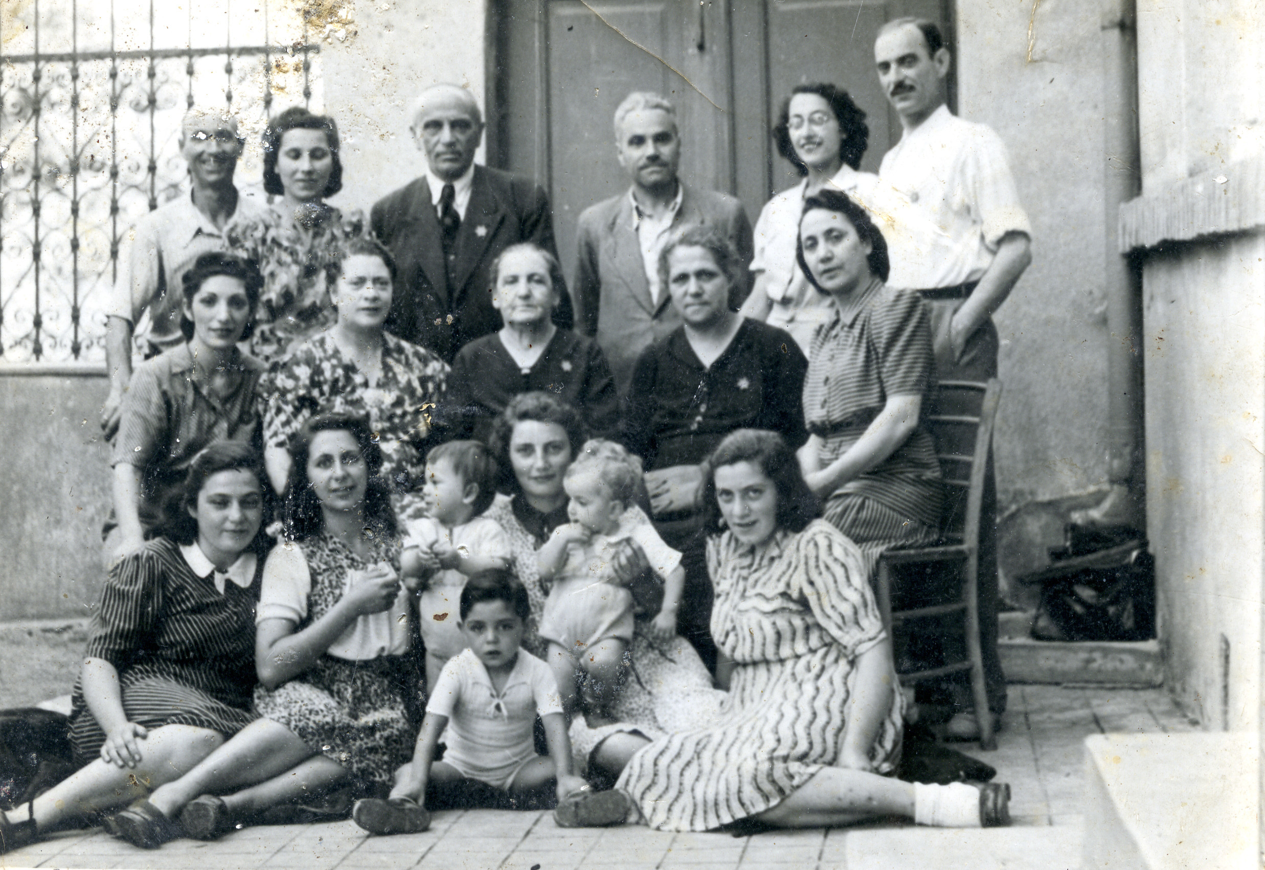 Group portrait of Jewish family members and neighbors after their expulsion from Sofia, in front of their house on Yumach street in Haskovo.

Among those pictured is Lore Beracha (seated in front, second from the left). Seated in the second row is her mother, Rashel (second from the right), and her best friend Lucy (far left).  The landlord, Abraham Yago, is standing in the back row, third from the right; his wife Mrs Yago is seated in the middle row, far right.  The other people pictured are neighbors.