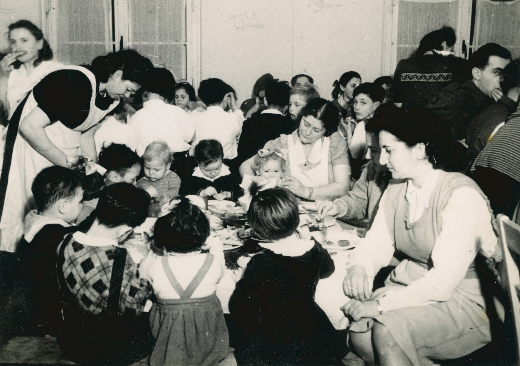Female aid workers feed young children in the Bergen Belsen displaced persons camp.

Pictured are Elana Millman (the blond baby in the center wearing a sweater) and Zippy Orlin, the aid worker sitting in front.