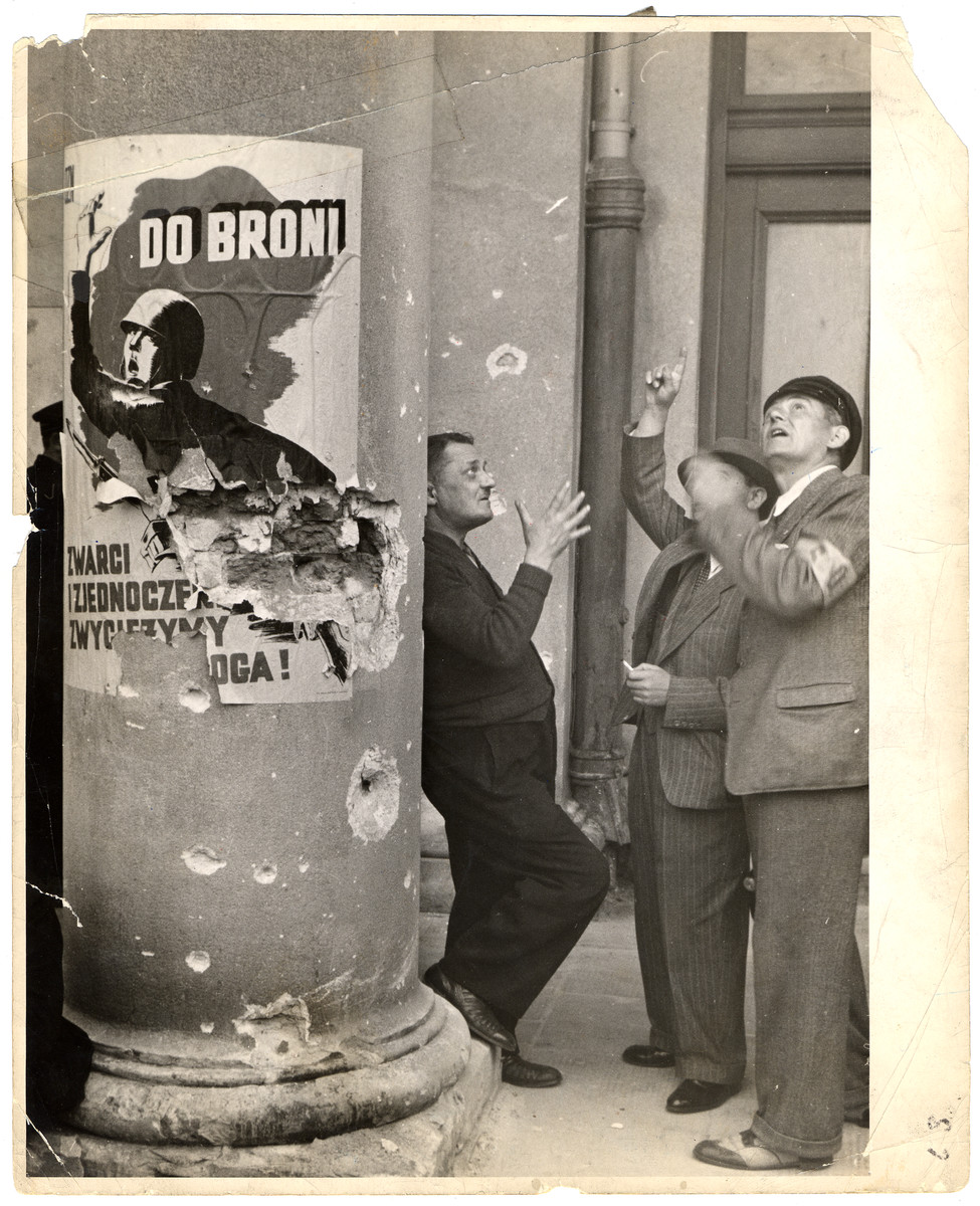 Three volunteer policemen (strag obywatelska) stand outside the opera house in besieged Warsaw. On the column next to the men is a propaganda poster that says: "To Arms. United We Will Defeat the Enemy!"