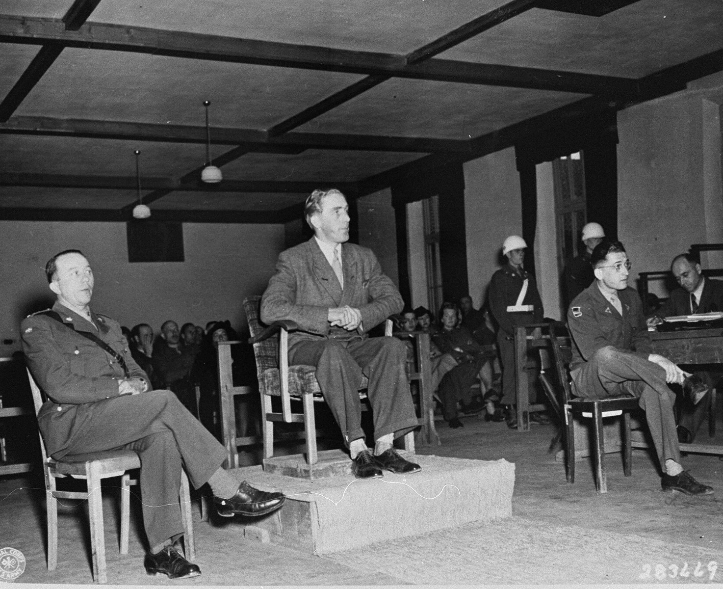 Aksel Frydenabjerg, a member of the Criminal Intelligence Division in Denmark, testifies at the trial of former camp personnel and prisoners from Buchenwald.  To his left is Captain Regner P. Mortensen, a Danish liaison officer and interpreter, and on the right is Herbet Rosenstock, another interpreter.