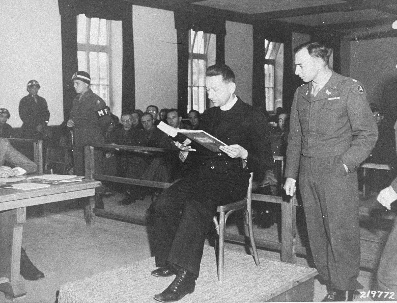 Polish priest Theodore Korcz reads to Lt. Colonel William Denson from a camp medical record presented as evidence at the trial of former camp personnel and prisoners from Dachau.  

The log recorded the deaths of several Catholic priests who were used as subjects in malaria experiments.