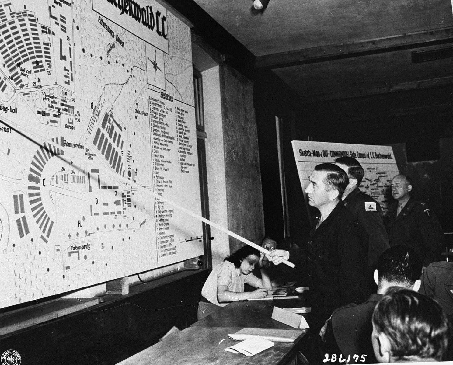 Defendant Josef Kestel indicates on a map where he shot a prisoner trying to escape, during the trial of former camp personnel and prisoners from Buchenwald.  

He is flanked on his right by Rudolph Nathanson, an interpreter, and Captain Groth, a defense attorney.