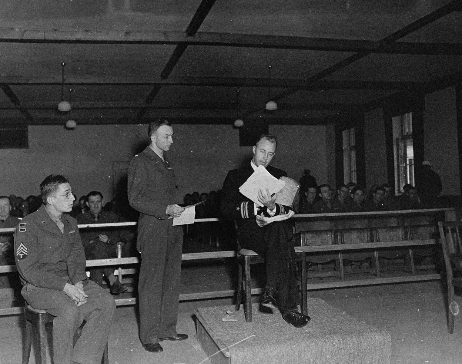 A witness testifies for the prosecution at the trial of 61 former camp personnel and prisoners from Mauthausen.  

Testifying is Lieutenant Jack Taylor (b. 1908).  Lt. Taylor served in the OSS (American Secret Service).  He was captured by the Germans and imprisoned in Mauthausen.  Also pictured are Herbert Rosenstock, an interpreter (far left) and U.S. prosecutor William Denson (middle).