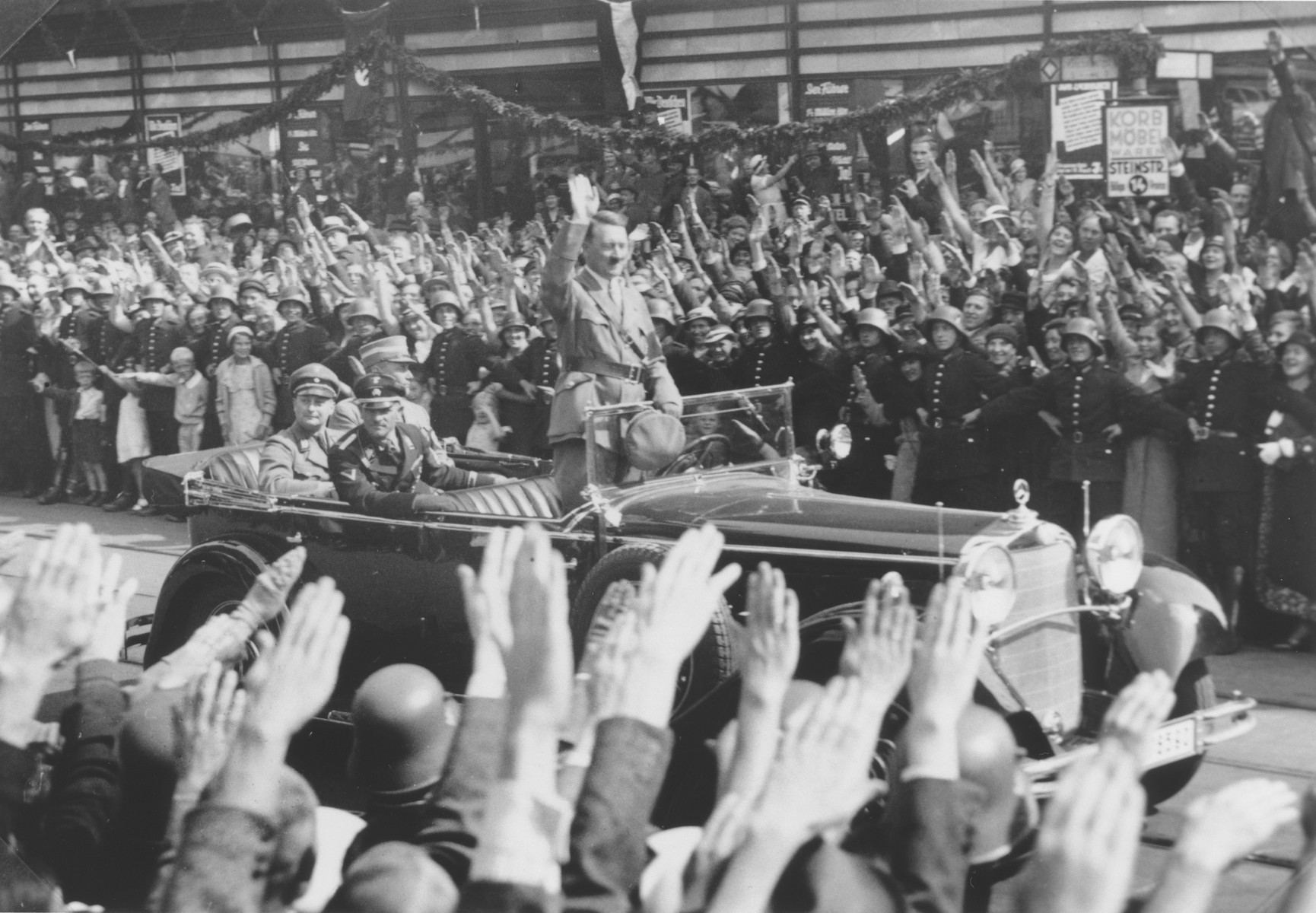 Standing in an open car, Adolf Hitler salutes a crowd lining the streets of Hamburg.

Pictured in the car with Hitler are the Gauleiter of Hamburg, Karl Kauffmann, and the SS-Obergruppenfuhrer Joseph "Sepp" Dietrich.  

One image from a photograph album belonging to a member of Police Battalion 101.