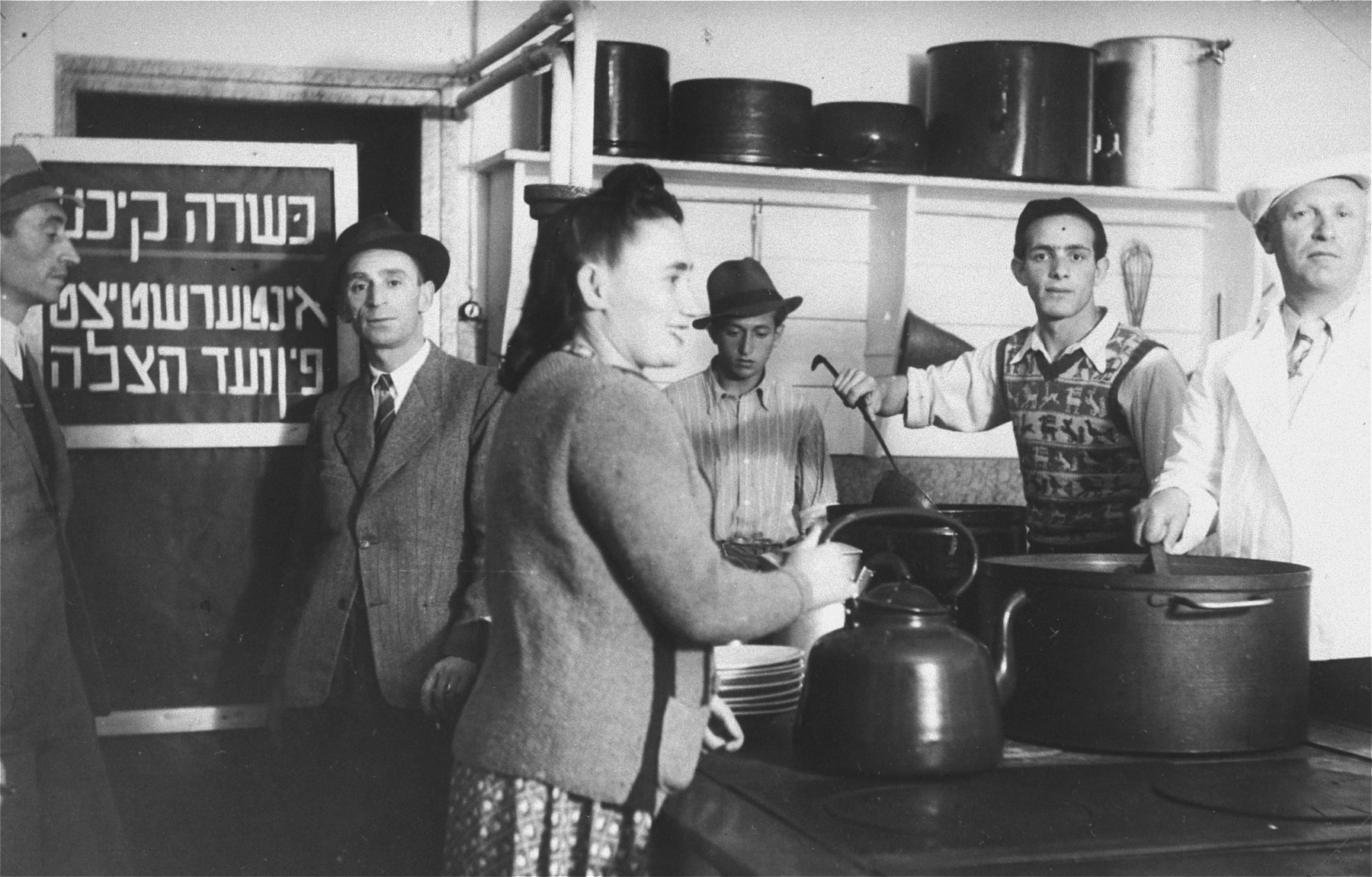 Jewish DPs at work in the kosher kitchen at the Mittenwald displaced persons camp.  

The Yiddish sign on the door reads: "Kosher kitchen, underwritten by the Vaad Hatzala [Rescue Committee].