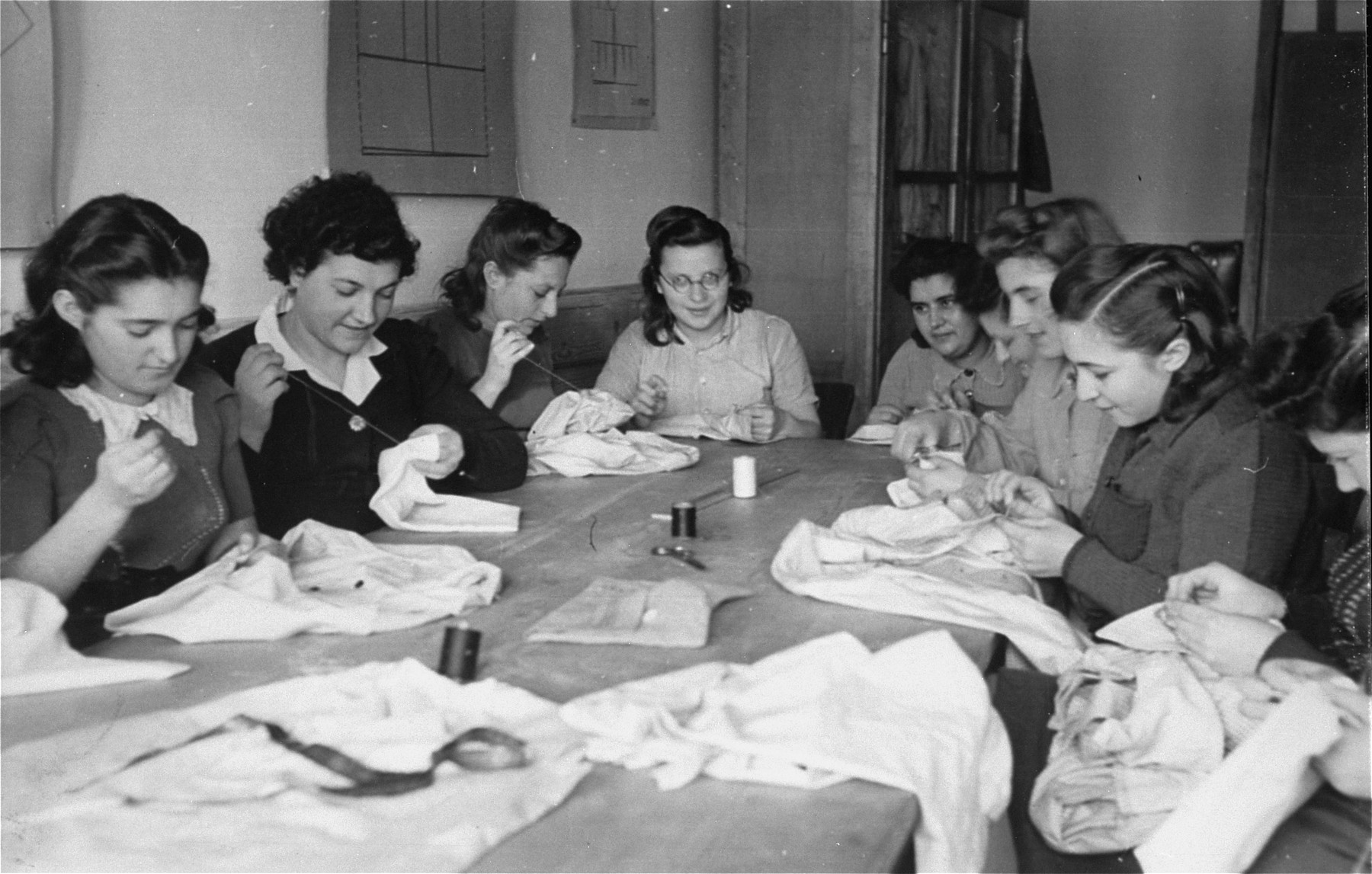 A group of women sit around a table and hand-sew garments in an ORT (Organization for Rehabilitation through Training) workshop in the Landsberg displaced persons' camp. 

Yiddish caption on the back: "Pioneer women of different kibbutzim (locations in Europe), learning to sew in ORT school". [Oversized print]