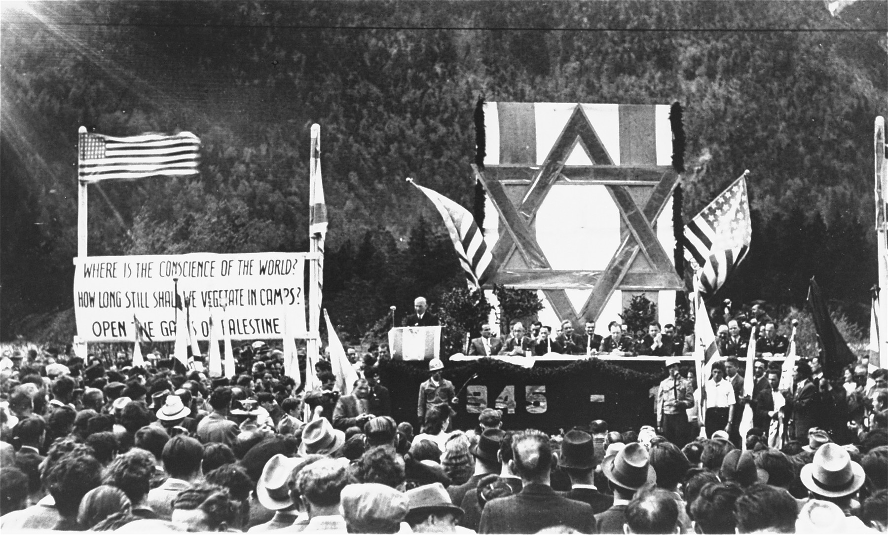 David Treger, President of the Central Committee of the Liberated Jews in Bavaria, delivers an address at a public meeting held in the Mittenwald displaced persons camp to protest British immigration policy in Palestine.  

The banners call for free immigration to Palestine.