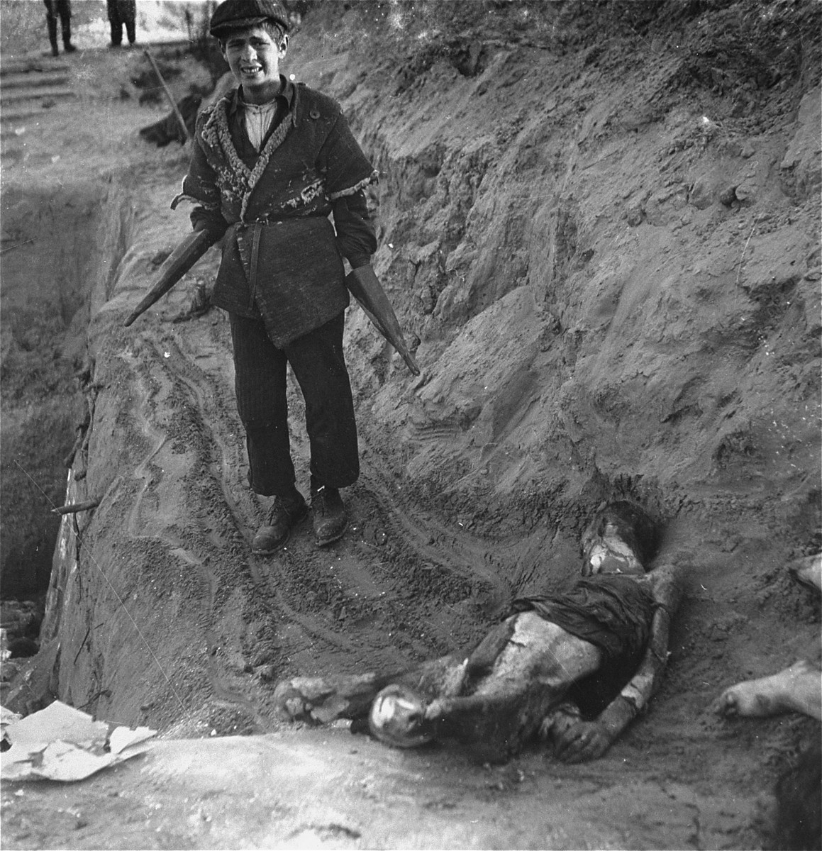 A boy at work burying bodies in the Warsaw ghetto cemetery pauses for Heinrich Joest to photograph him.  

Joest's caption reads: "Himself still half a child, this youth belonged to the Jewish corpse-bearers: he showed me how he took hold of the corpses with his remarkable gloves, pulled them to the edge of the grave and let them slide down."