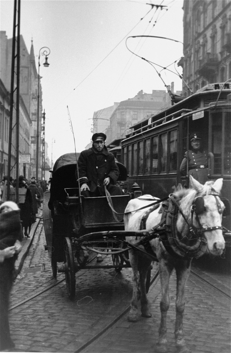 A driver leads a horse-drawn carriage along a street in the Warsaw ghetto.

Two German police officers are visible on the streetcar at right.