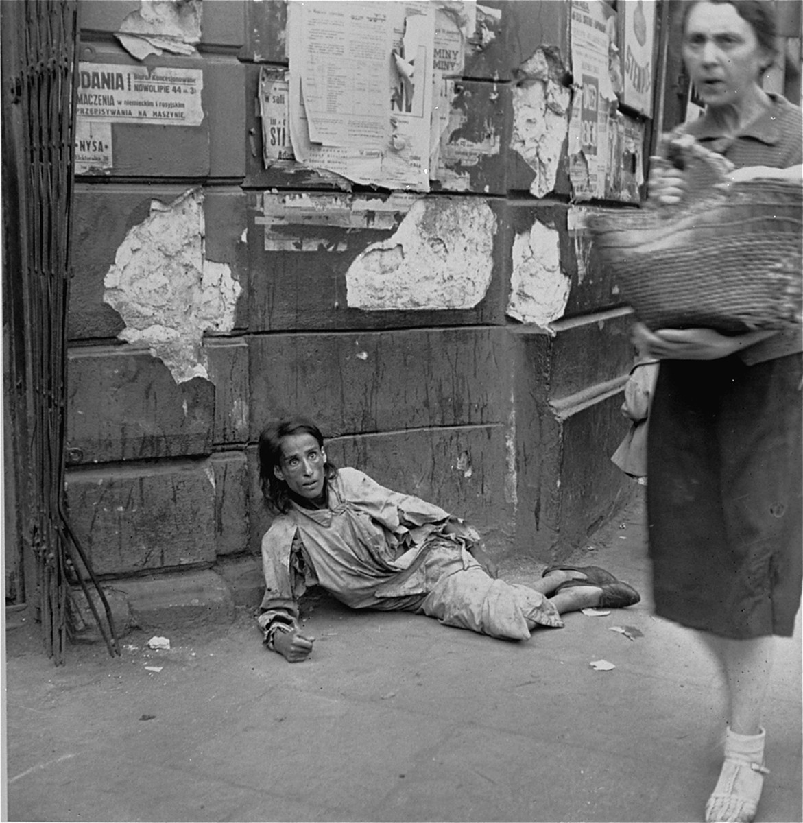 A destitute woman lies on a sidewalk along Nowolipie Street in the Warsaw ghetto.  

Joest's caption reads: "I photographed this begging woman with fixed, staring eyes on Nowolipie Street as people went by.  [Hanging] above her was a small placard advertising an office on 44 Nowolipie Street where German and Russian could be translated."
