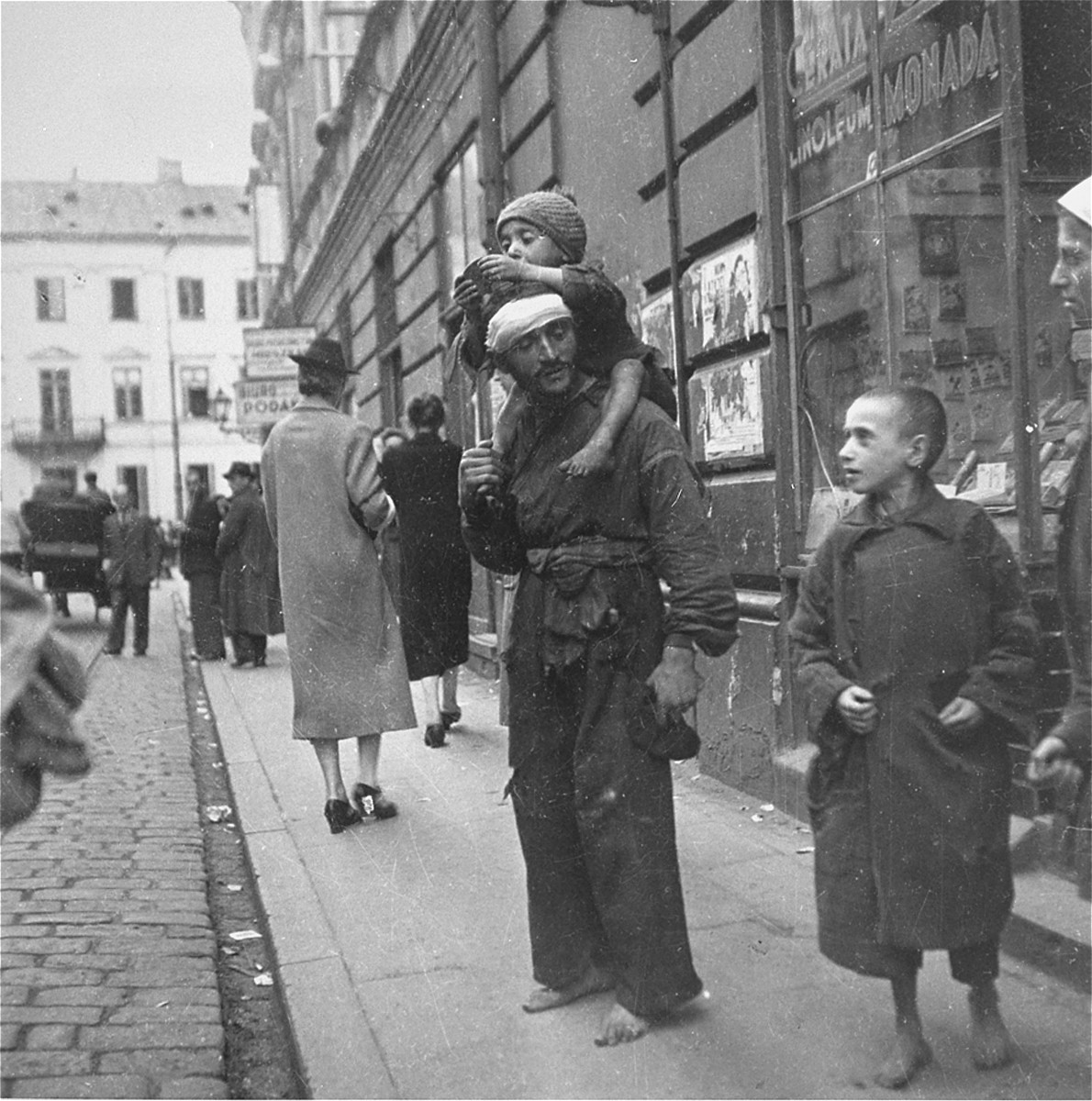 A destitute Jewish man with his two children on a street in the Warsaw ghetto. 

Joest's original caption reads: "Next to a lady with a good coat and shoes that had heels, stood next to this broken-down man, barefoot, with his barefoot child on his shoulders."