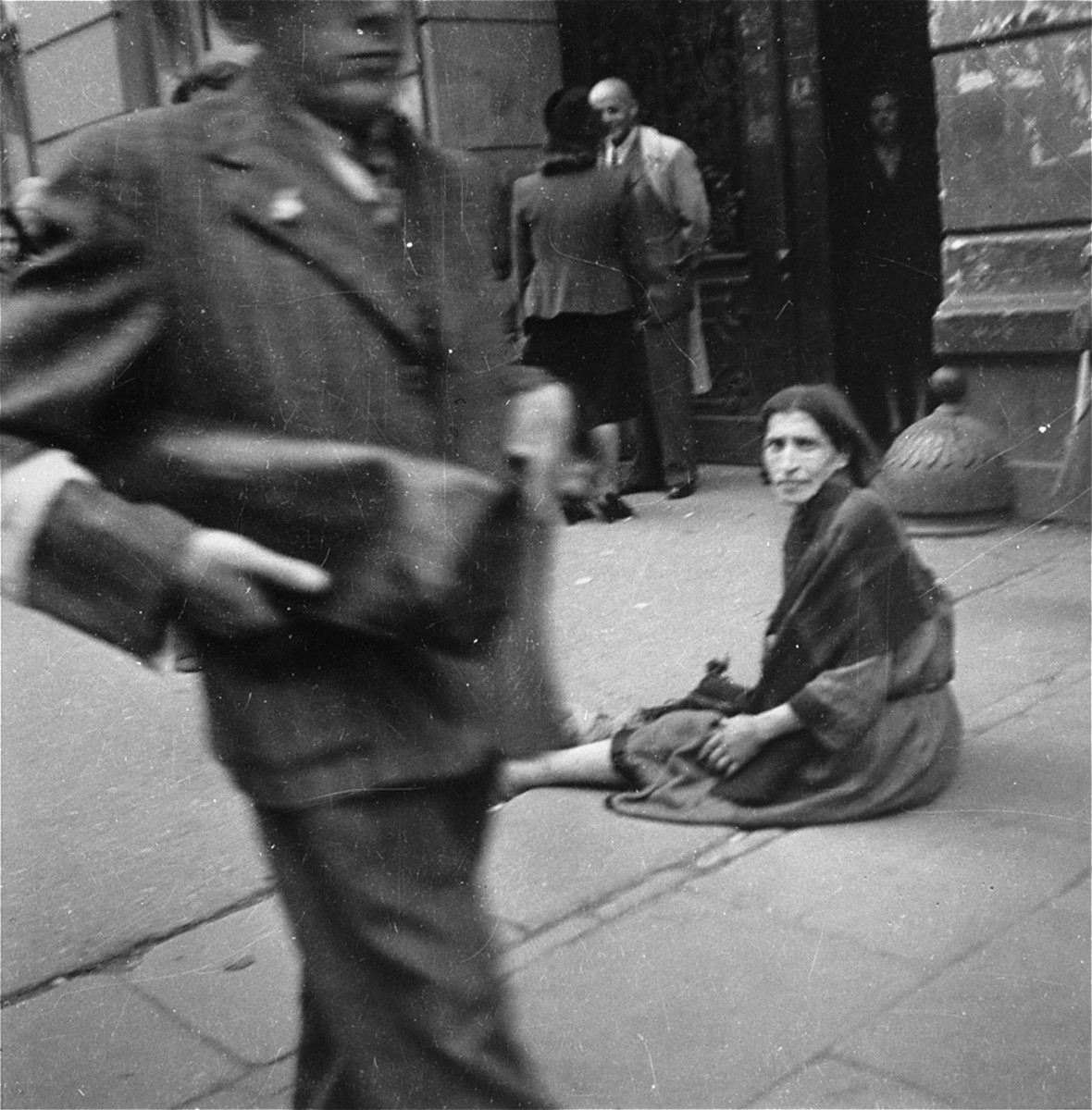 A destitute woman sits in the middle of a busy sidewalk in the Warsaw ghetto.  

Joest's caption reads: "In the middle of the sidewalk sat this wailing woman, over whom no one troubled himself, and begged.  What was astounding was that from time to time she received something even though everything was going so badly with all the others as well."