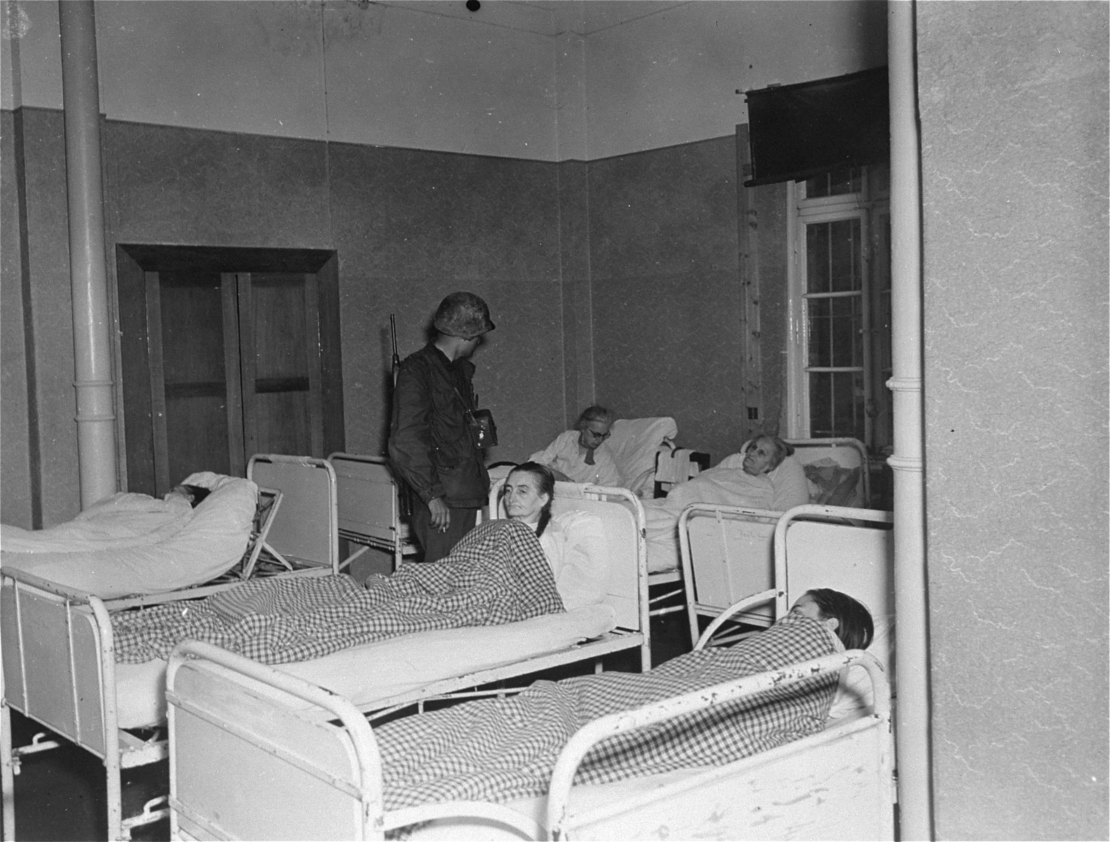 Lt. Alexander J. Wedderburn, photographer with the 28th Infantry Division, First US.Army, questions elderly survivors who are lying in bed at the Hadamar Institute. 

The photograph was taken by an American military photographer soon after the liberation.
