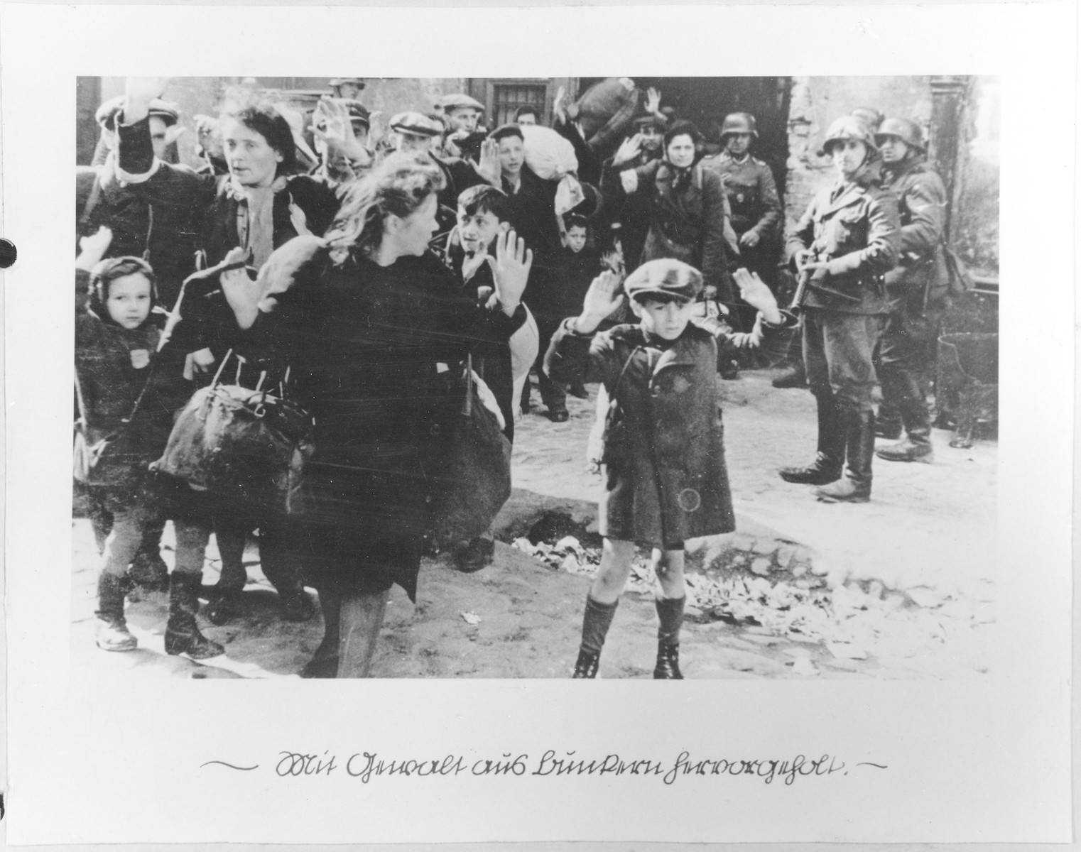Jews captured by SS and SD troops during the suppression of the Warsaw ghetto uprising are forced to leave their shelter and march to the Umschlagplatz for deportation.  

The original German caption reads: "Pulled from the bunkers by force."

The SD trooper pictured second from the right, is SS-Rottenfuehrer Josef Bloesche, who was identified by authorities using this photograph.  Bloesche was tried for war crimes by an East German court in 1969, sentenced to death and executed in July of that year.  

The little girl on the left has been identified as Hanka Lamet, who is standing next to her mother, Matylda Lamet Goldfinger (the woman second from the left).  The boy carrying the sack has been identified as Leo Kartuzinsky and the woman in the front has been identified as Chana Zeilinwarger.  Numerous people have identified the boy in the foreground as either Arthur Domb Semiontek, Issrael Rondel, Tsvi Nussbaum or Levi Zeilinwarger, but none of these identifications can be conclusively corroborated
