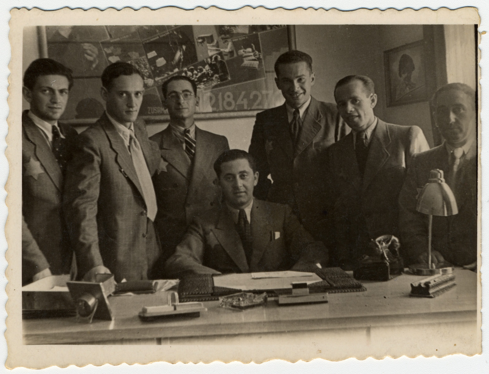 Group portrait of employees of the leather workshop in the Lodz ghetto standing in front of a large photographic collage.

Among those pictured is Leon Fajtlowicz (standing third from left).  He was the uncle of the donor and in charge of all the leather workshops in the ghetto.