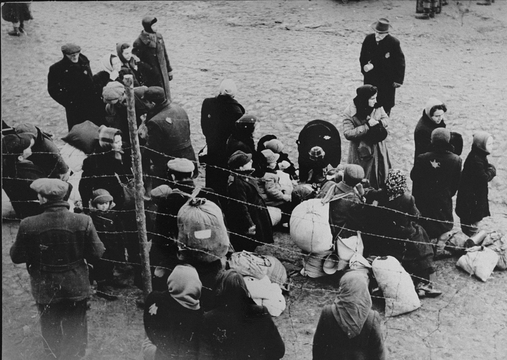 A group of Jews is gathered at an assembly point in the Kovno ghetto during a deportation action. 

[Though the photo seems to be part of the series of the deportation to Estonia, the family has been identified as the Ipp family who were sent to Riga.  Among those pictured may be Jacob Ipp (now Ipson, the boy standing on the left, behind the barbed wire fence), his mother, Etta (Butrimowitz) Ipp (standing behind him), and Chananya Butrimowitz (Etta's father, carrying a backpack).]