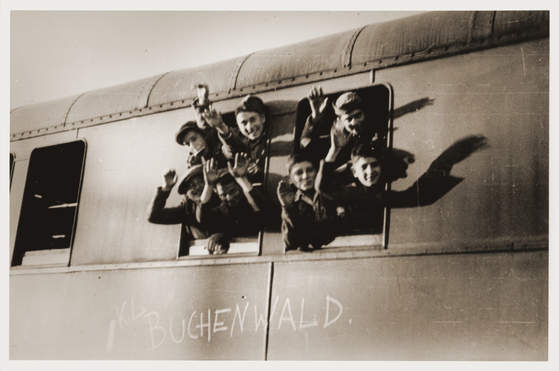 Jewish youth liberated at Buchenwald wave from the window of a train, as it pulls away from the station.  

The train, which has been marked with the words "KL Buchenwald," will transport the children to an OSE (Oeuvre de Secours aux Enfants) home in Ecouis, France.  Pictured in the upper right hand corner is Leon Friedmann.