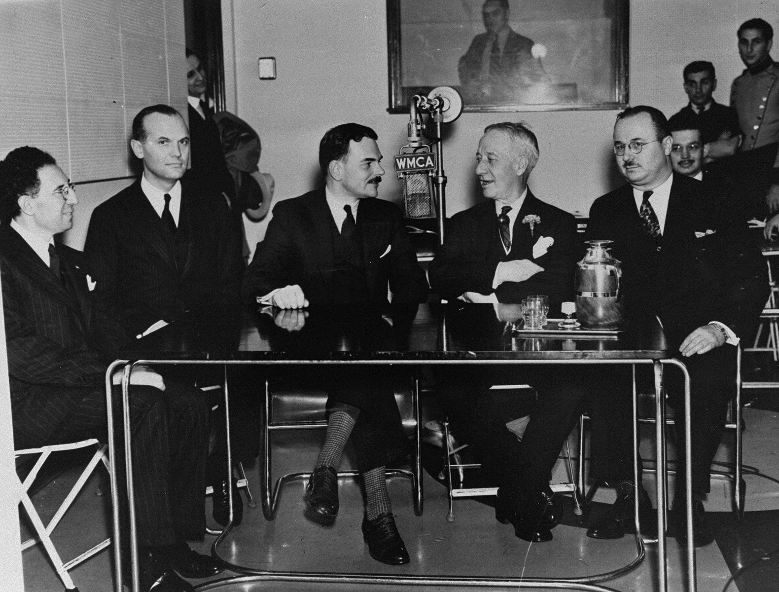 Five public figures sit around a table in the studios of radio station WMCA, where they will participate in a radio broadcast condemning the Nazi persecution of German Jews.  

Pictured from left to right are: Johan Smertenko, Executive Director of the Non-Sectarian Anti-Nazi League; the Reverend Elmer McKee, Rector of St. George P.E. Church; District Attorney Thomas E. Dewey; Former Governor Alfred E. Smith; and Dr. Mitchell Salem Fisher, a member of the Anti-Nazi League.