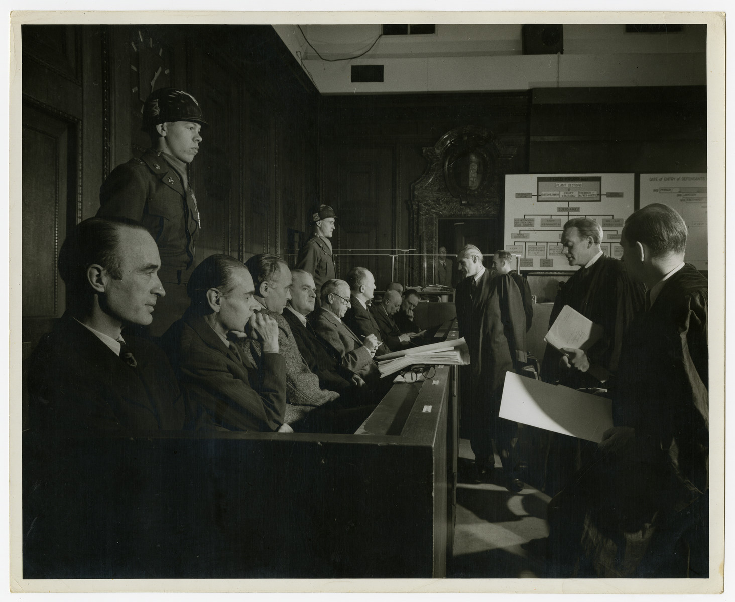 Close-up view of the defendants of the Krupp case.  Behind them are military police.

Seated in the front left is the chief defendant Alfred Krupp von Bohlen.  In the back is a poster showing organizational structures.