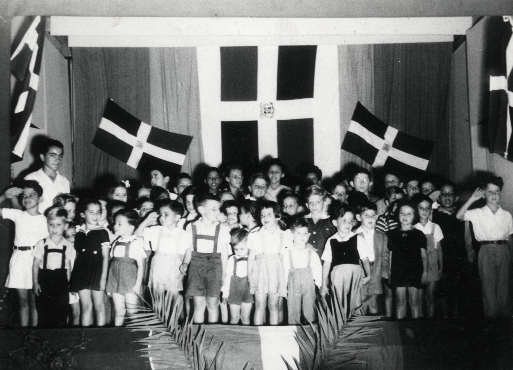 Young children stand in front of Dominican flags at a ceremony in their school in Sosua.  

Teenager Marcel Salomon stands behind them on the left.