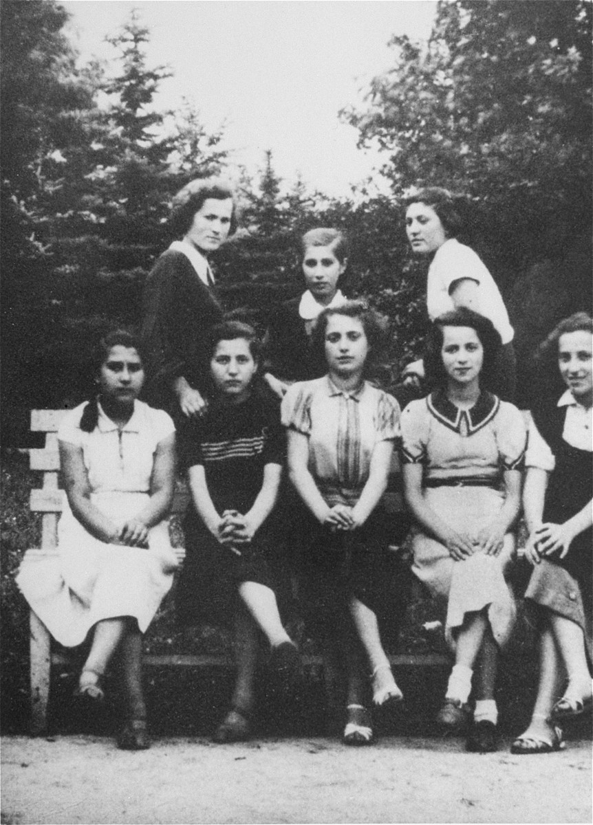 A group of teenage girls who are members of the Hashomer Hatzair Zionist youth movement, pose on a park bench in Piotrkow Trybunalski. 

Pictured sitting from left to right are: Hajka Zomper, Bella, Rozia Cwiling, Genia Goldszmidt and Pola Pudlowska. Standing from left to right are: Rozia Gomolinska, Cipora Cwiling and Salla Landau.