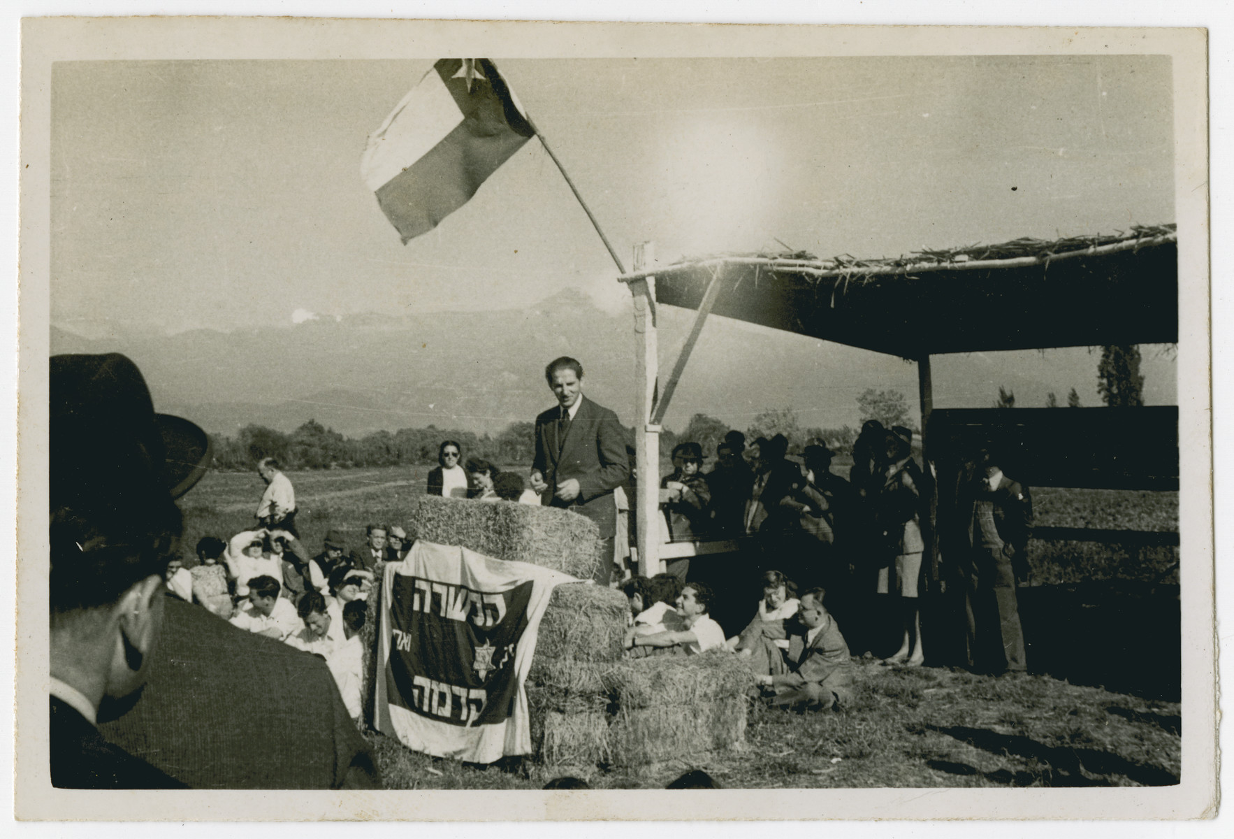 Photograph from an album entitled, "Hacshara Kidma Chile,"  documenting life on a postwar Shomer Hatzair Zionist agricultural collective in Chile.

A man addresses the crowd from a strawbale podium, under the Chilean flag.  The inscription on the back reads, "Jose Tchormitzki Meseico."  The Hebrew banner says Hachshara Kedma.