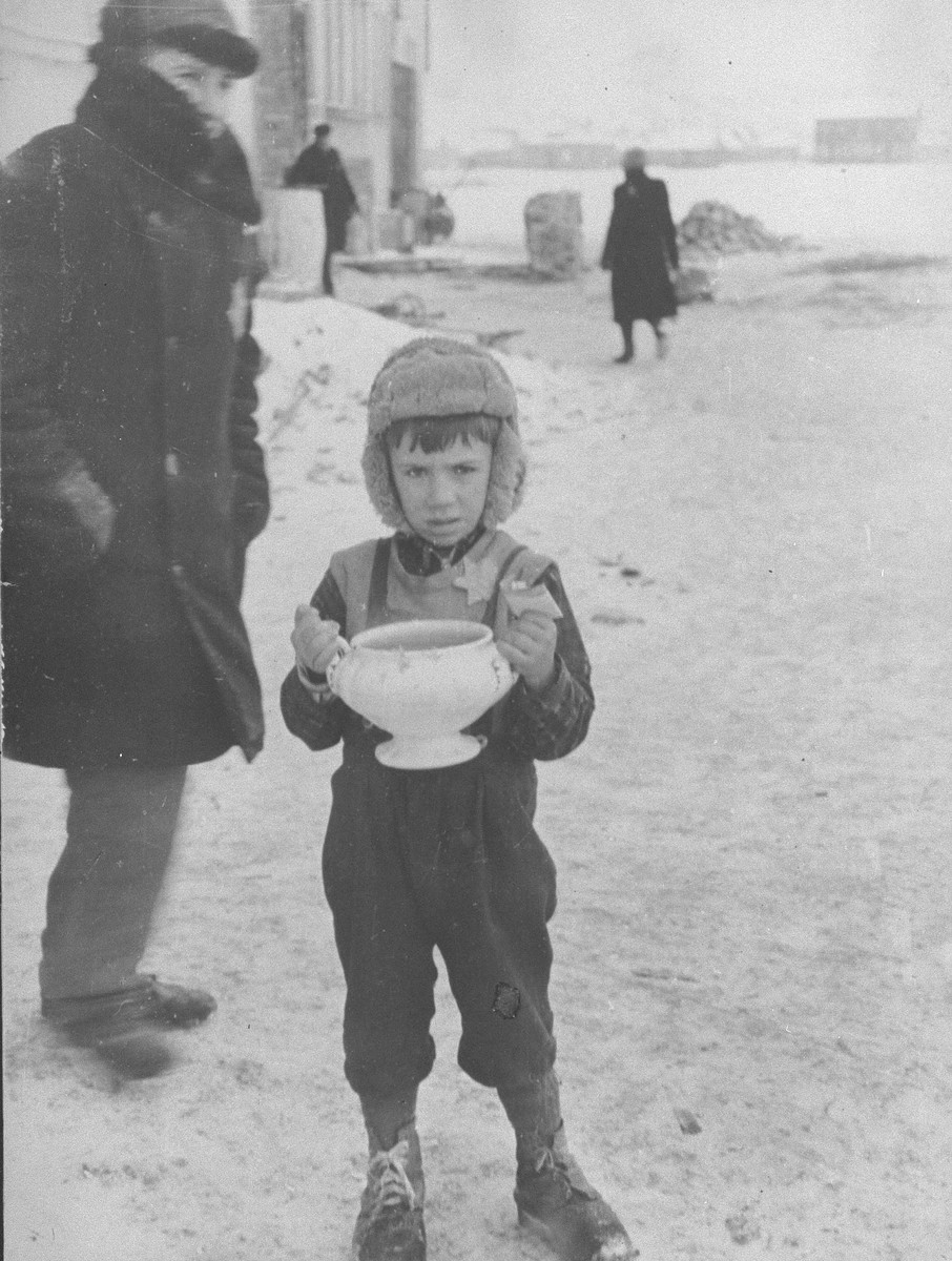 A young boy in the Kovno Ghetto carrying a bowl of soup and holding food ration tickets.

According to Solly Ganor "A five-year-old boy receives hot soup for the family from the ghetto soup kitchen. His father was murdered in the beginning of the war and the mother had to take care for the smaller children. The boy became the provider of the family. I saw him often standing at the corner trying to sell what was left of the family possessions. He was shot trying to escape from the Germans during the "Children's Action" on March 27, 1944. "