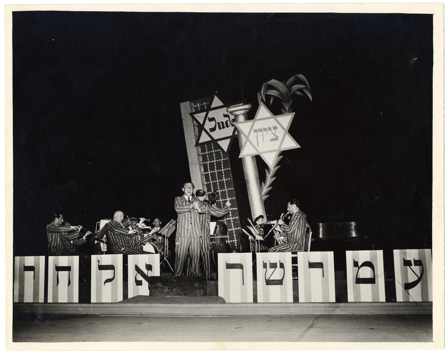 The Saint Ottilien Ex-Concentration Camp Orchestra performs a concert in Munich for American soldiers and UJA staff.

The sign in front reads "Am Yisrael Chai" (the nation of Israel lives).  

Pictured from left to right are Max Beker (violin), Max Borstein (violin), Melech Granat (drums), Jerzy Richter (voice), Fania Beker (piano) and Rala Wolfberg (clarinet).

See photograph 29734  for alternate caption.