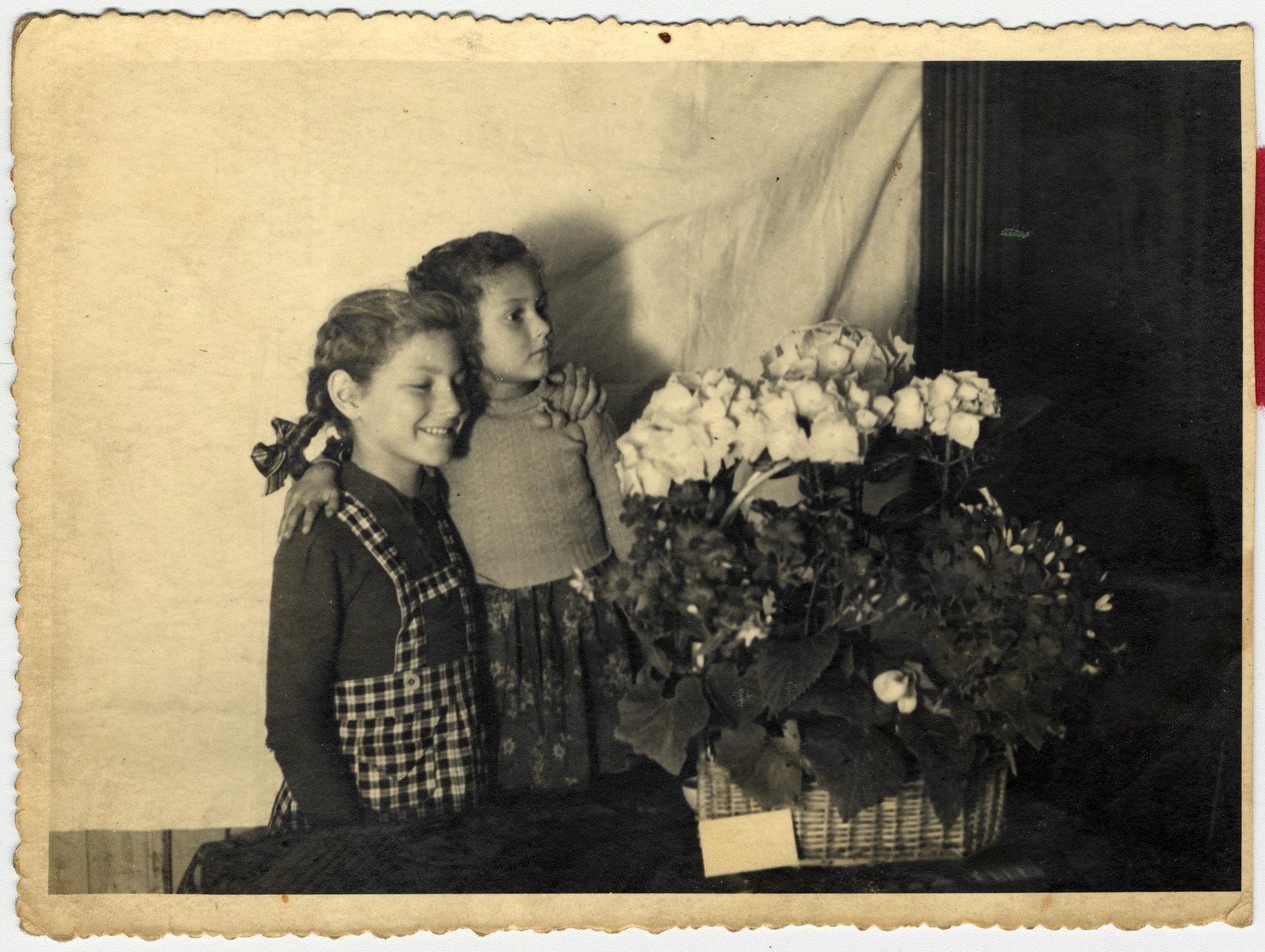 Sisters Halina  and Eva Litman pose next to a basket of flowers while in hiding in Jaroslaw. 

Eva is standing on a chair so they appear to be the same height.