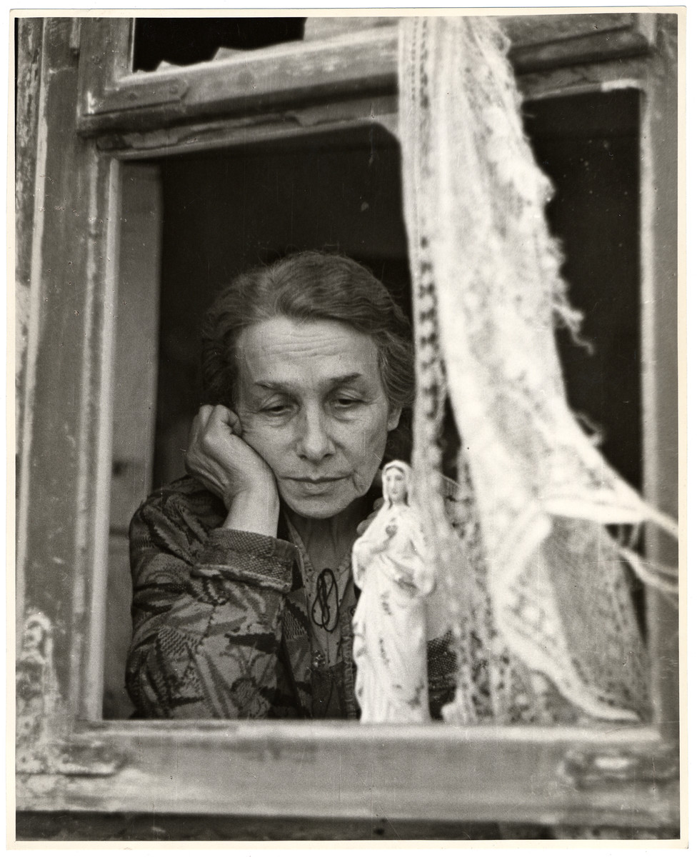 An elderly Polish woman looks out the shattered window of her apartment in Warsaw, where the remnant of a lace curtain hangs next to a statue of the Virgin Mary.