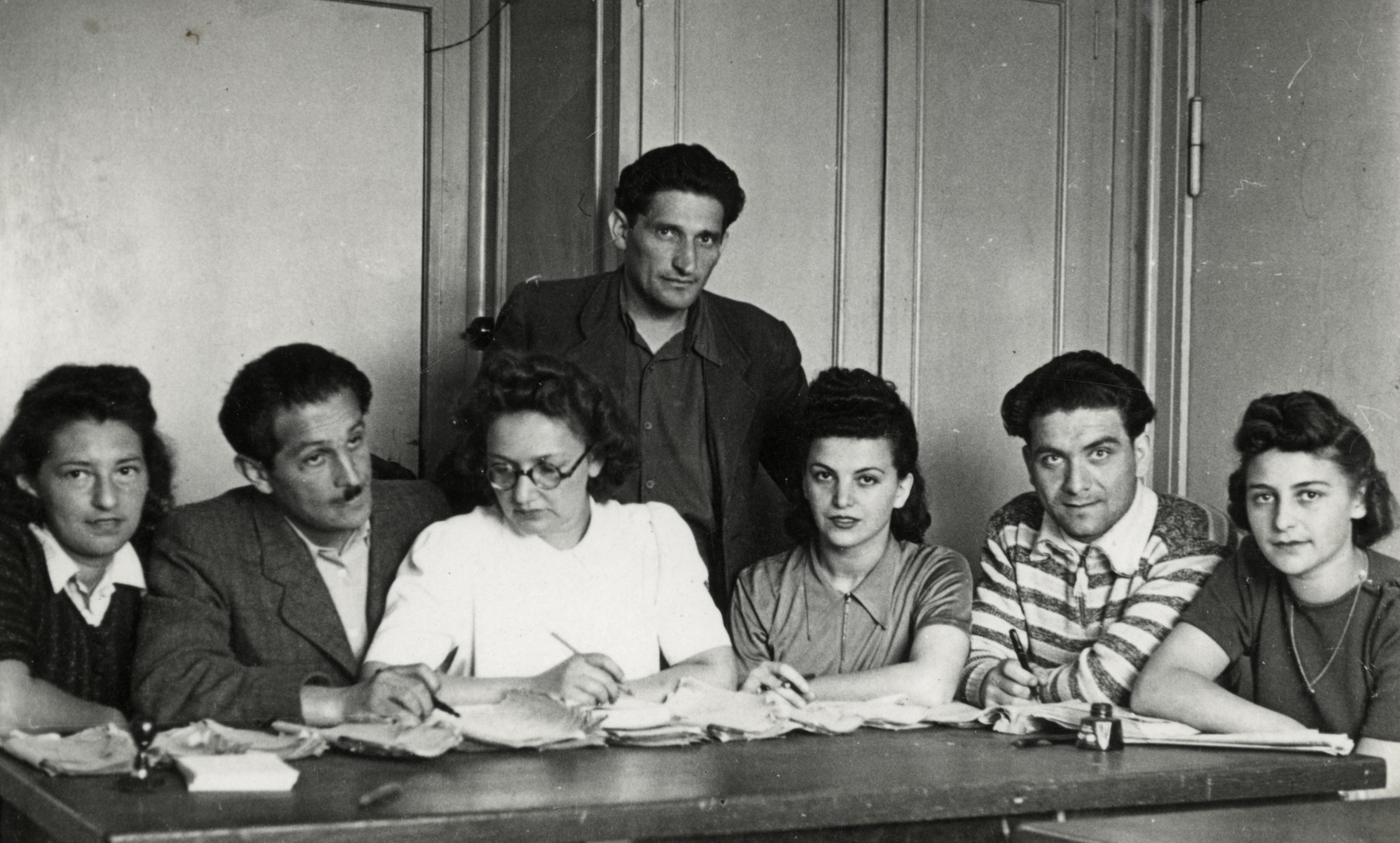 Office workers in the Schlactensee displaced persons camp work at a table. 

Among those pictured are Ms. Langsam (third from left), Mary Binder (third from right), and Rozalia (Krysia Laks) Lerman (far right).  Tuvia Grossman has also been identified in the front right.