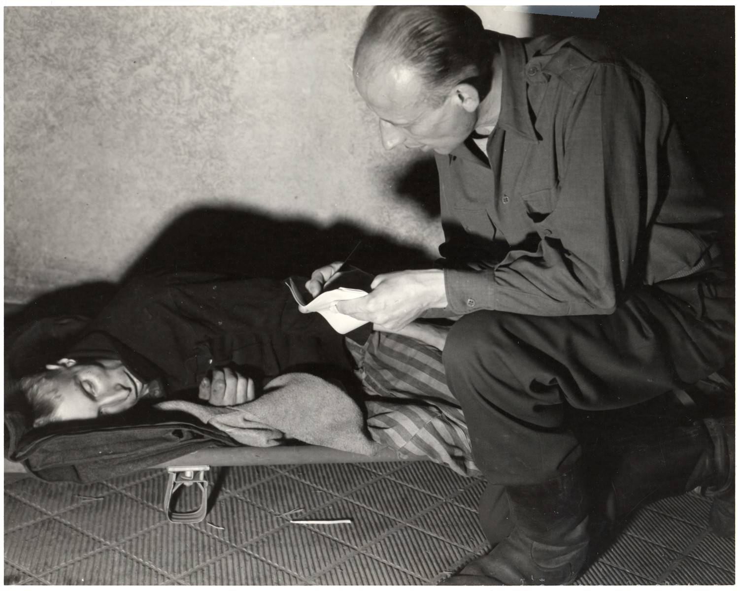 A man kneels next to an enfeebled survivor of the Woebbelin concentration camp and takes notes.