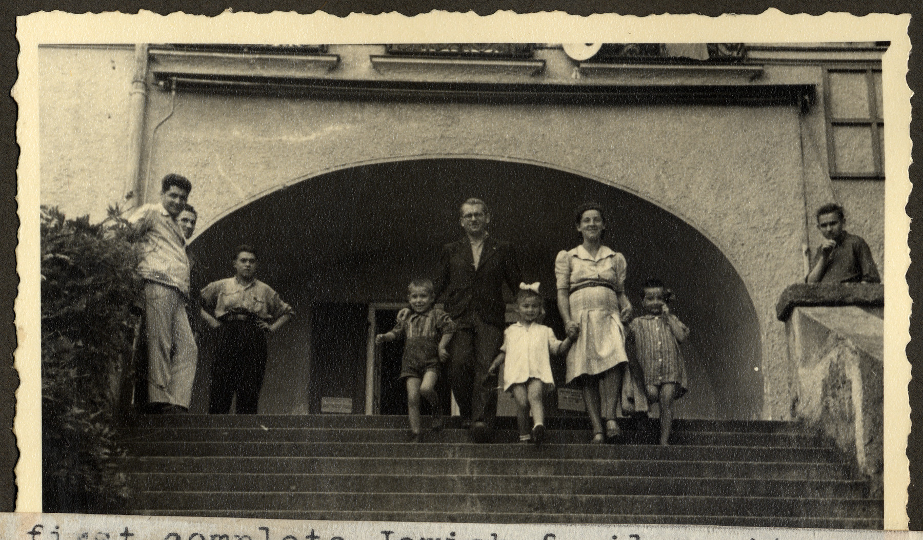 A reunited Jewish family walks down an exterior  flight of stairs in the Feldafing displaced persons' camp.

The original caption reads: "The first complete Jewish family unit from Poland.  Parents survived years in concentration camps; their little girl was hidden with Polish peasants, until the parents called for her.  The two boys are orphans, -- only other children at Feldafing."