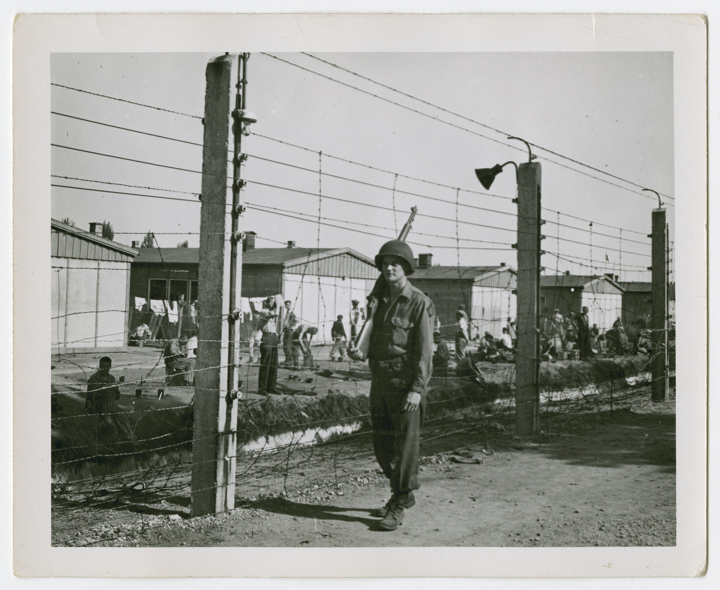 An American soldier guards the perimeter of the Dachau concentration camp.  Behind the fence are either survivors or captured Nazi war criminals.