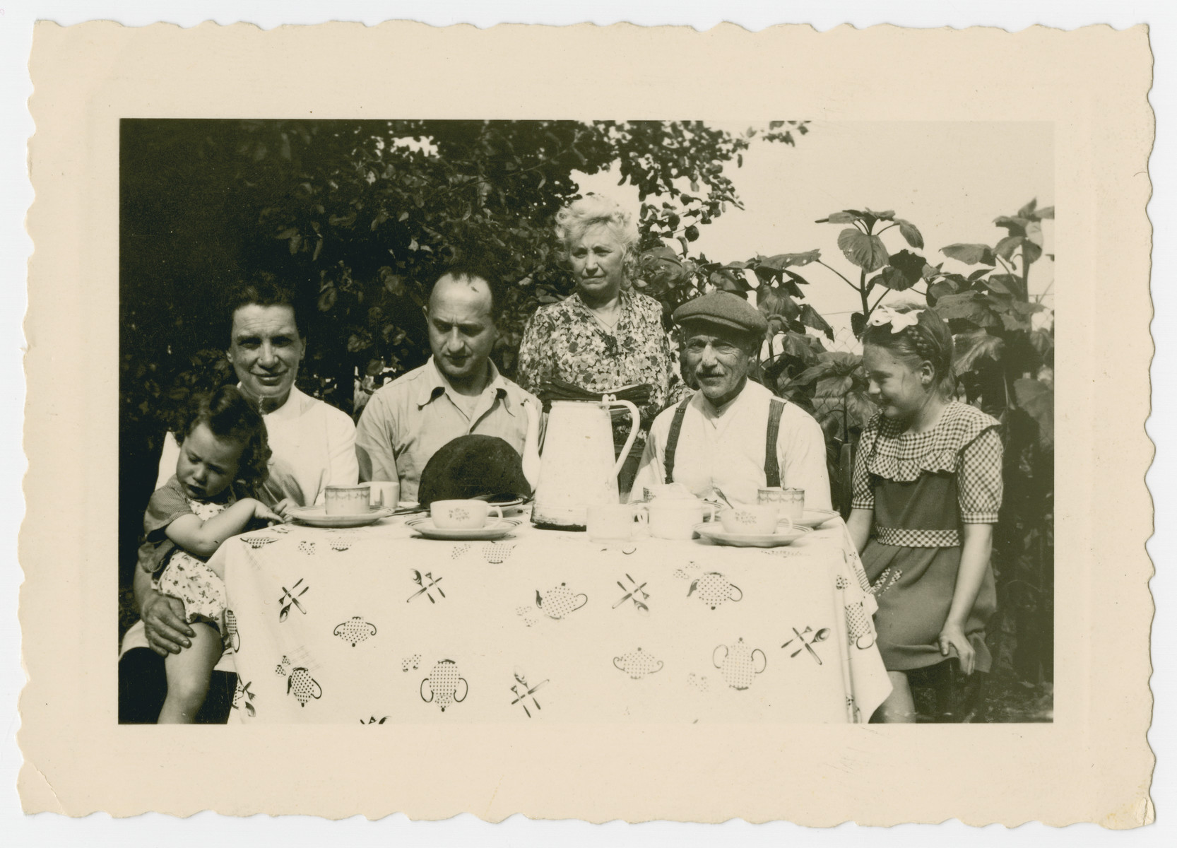 Shirley Gola sits at a table with her rescuers and family while in hiding.

Shirley Gola is seated on the lap of her rescuer Marie Fannes. Her father Joseph Gola is seated next to her.  Marrein, her godmother is standing next to her husband Monsieur Mal.  Their young niece is seated on the left.