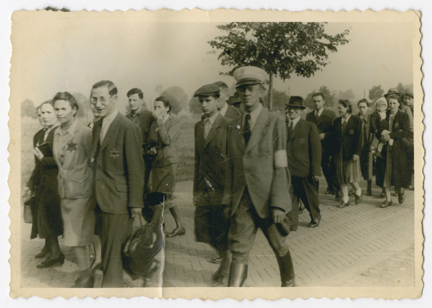Jews march down a street in Bedzin accmpanied by a Jewish policeman.

Henri and Esther Borzykowski (nee Fiszer) pictured in the back right walk in a line during their time in Bedzin. The two married before the war and survived the camps. Esther's father Srul Fiszer is pictured in front of the couple in a top hat.
