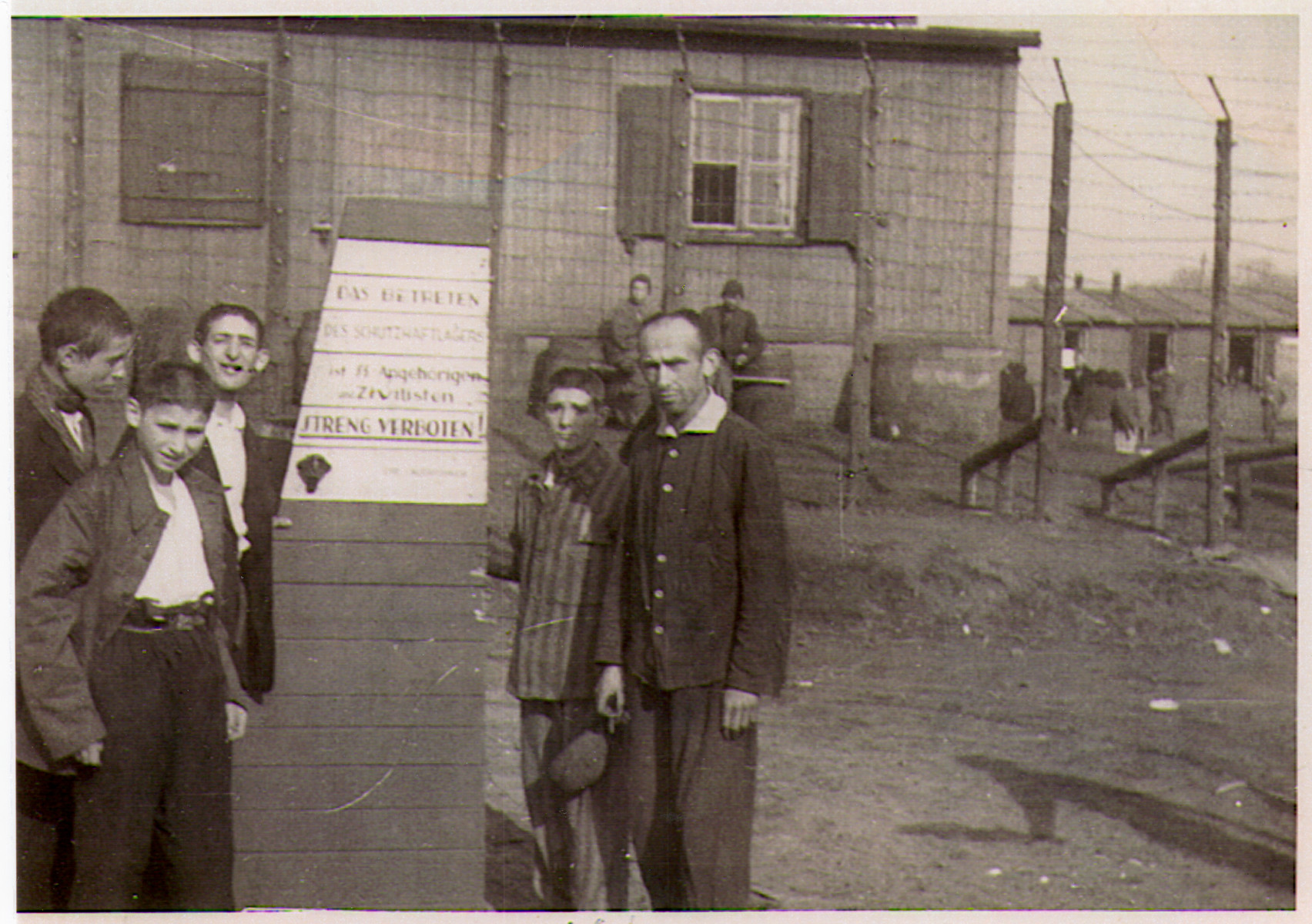A group of survivors, including many children, stand in front of the barbed wire fence at the entrance to the Hanover-Ahlem concentration camp.

The German sign posted in front of the fence reads "The entrance to the prison camp by relatives of the SS and civilians is strictly forbidden."