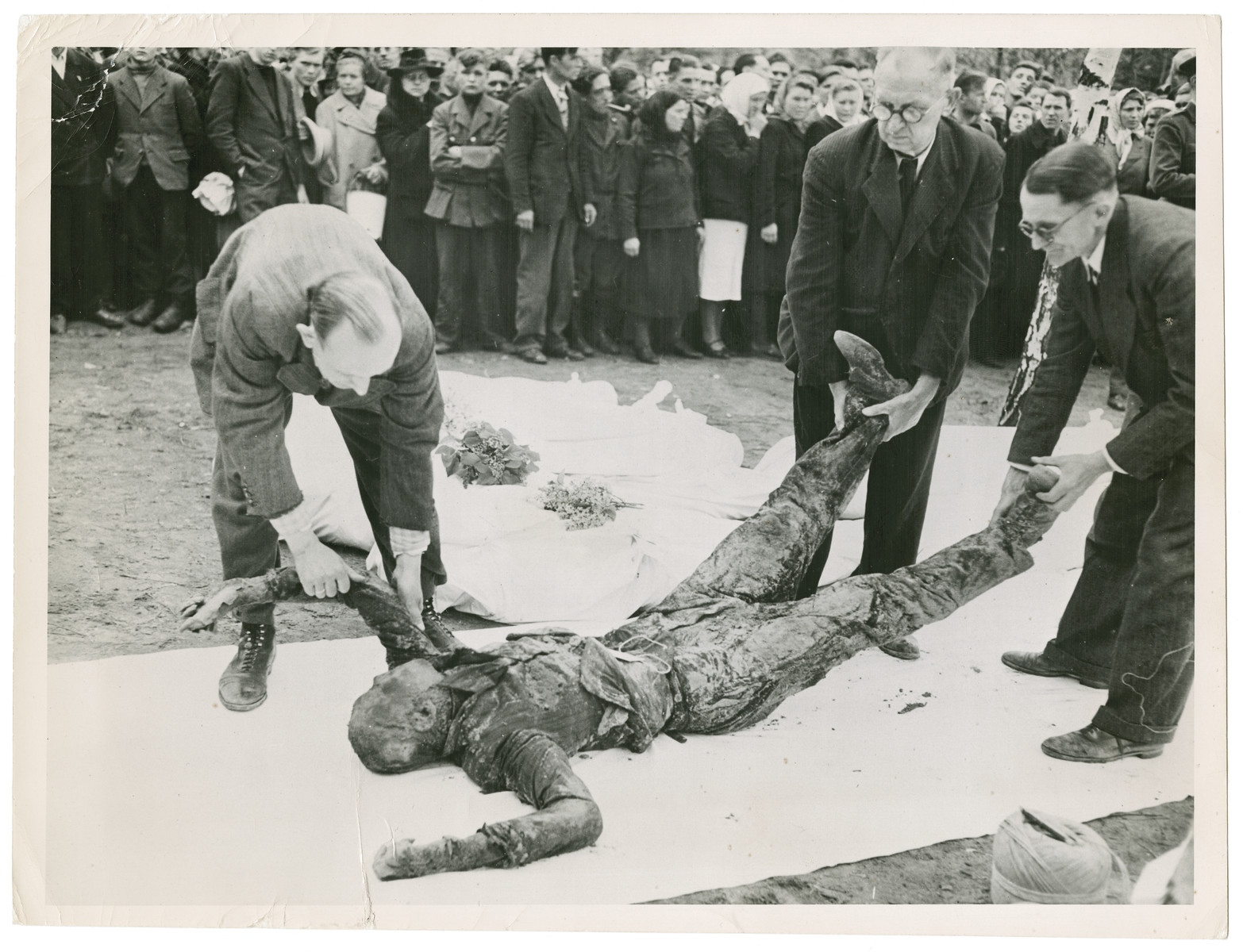 German civilians rebury the exhumed corpses of Russian prisoners.

Original caption reads:  "German civilians place the exhumed body of a Russian prisoner on a white sheet of decent burial.  The bodies of other victims lie enshrouded and covered with flowers in the background".