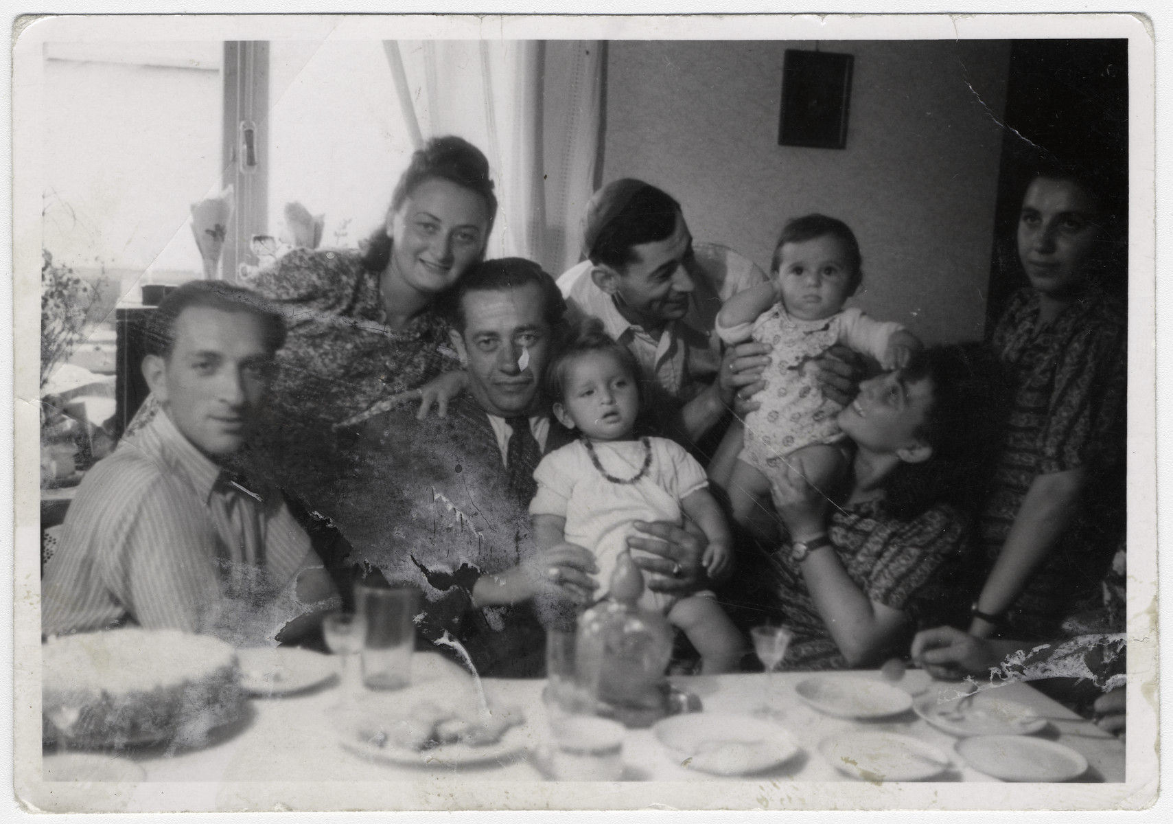Men and women gather around a table for a celebration in the Bergen-Belsen displaced persons camp.

Left to right (sitting): The second gentleman holding the baby is Elias Rosengarten