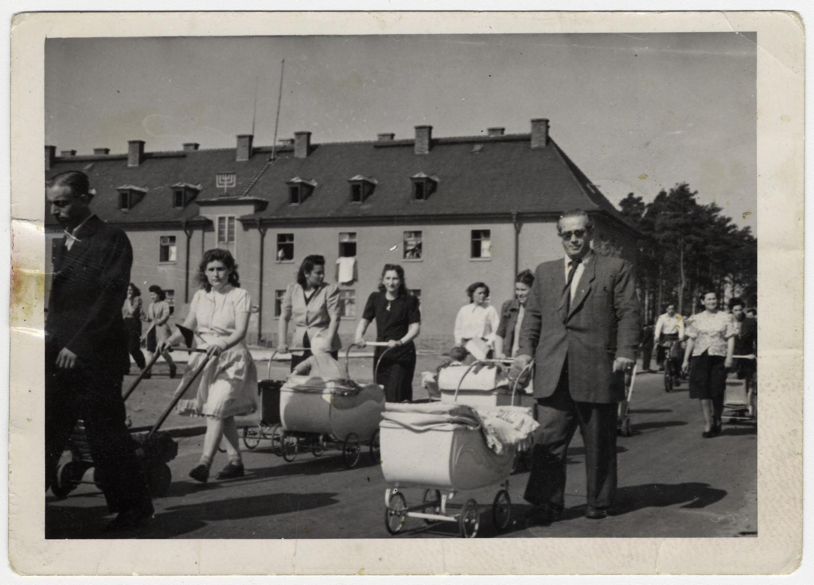 Men and women walk through the central plaza of the Bergen-Belsen displaced persons camp pushing baby carriages.

The gentleman on the right side wearing tinted spectacles, with his right hand on the stroller is Elias Rosengarten.