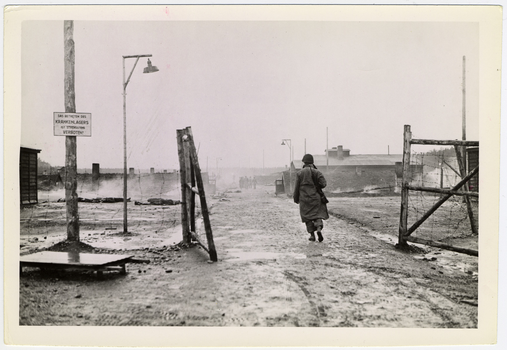 An American soldier follows others into the still smoldering ruins of the Landsberg (Kaufering Lager I) concentration camp, sub-camp of Dachau.

Original caption reads: "Landsberg atrocity: 7th U.S. Army troops enter the smoking ruins of the Landsberg concentration camp. Before American forces captured the camp, German guards locked their prisoners in the wooden huts and set fire to them. Some burned to death in the buildings, others found strength to crawl out and die in the streets. Most prisoners were naked, but some had remnants of clothing on their charred bodies."