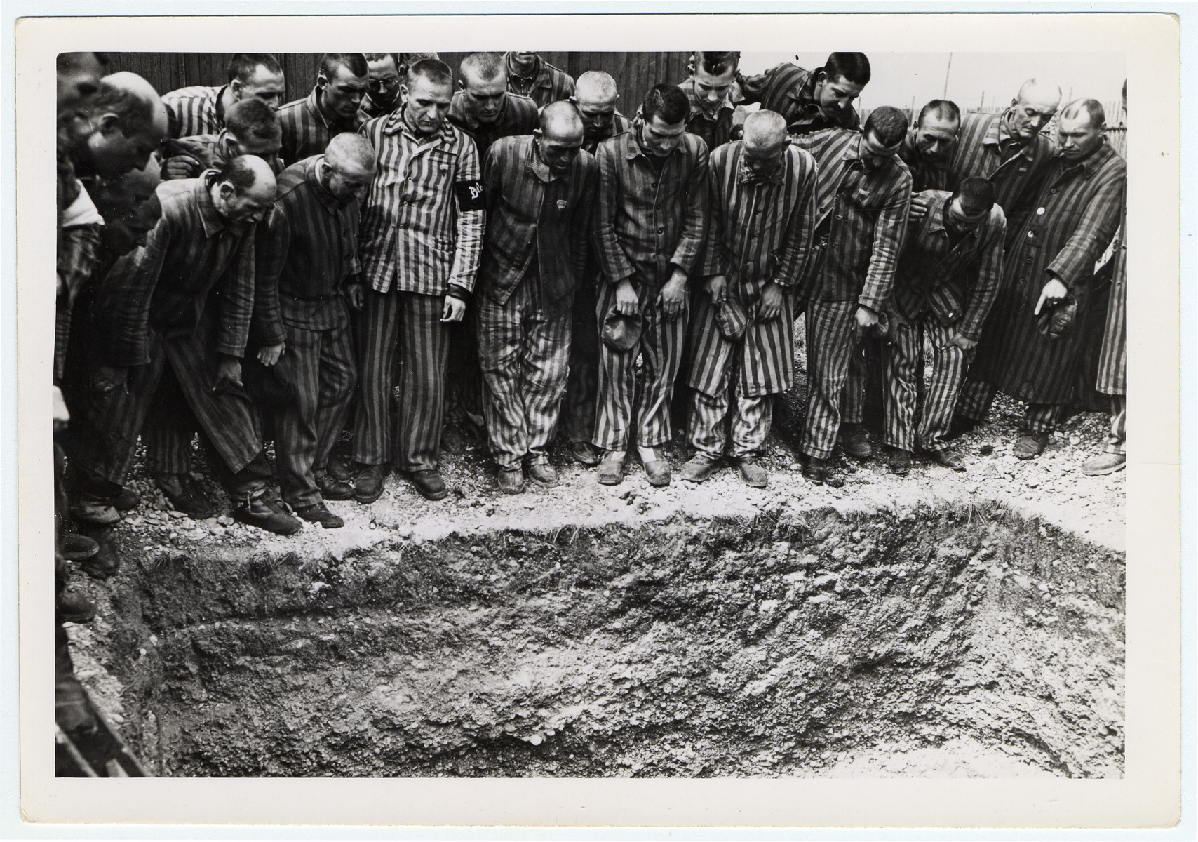 Newly liberated prisoners stand around a mass grave at Dachau concentration camp.

Original caption reads, "The prisoners show where they buried some of the comrades everyday, the ones who could not withstand the cruel treatment and starvation diet handed out by their German keepers. Feet and partially covered head protrude from under the dirt in this mass burial pit."