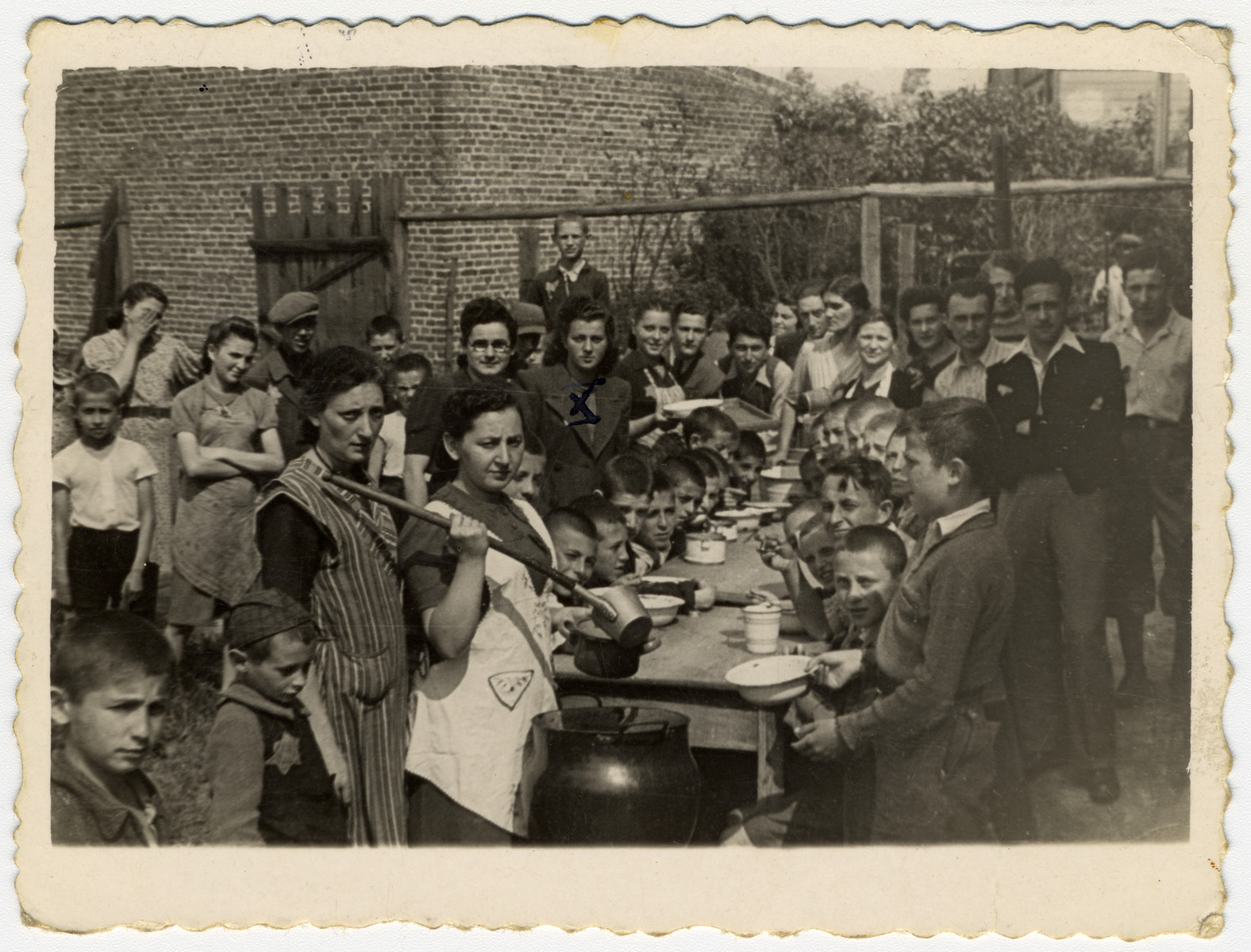 Children in an orphanage in Marysin sit around an outdoor table awaiting the distribution of soup. 

In the middle, dividing the food, is the director of the children's home, Mrs. Litman.