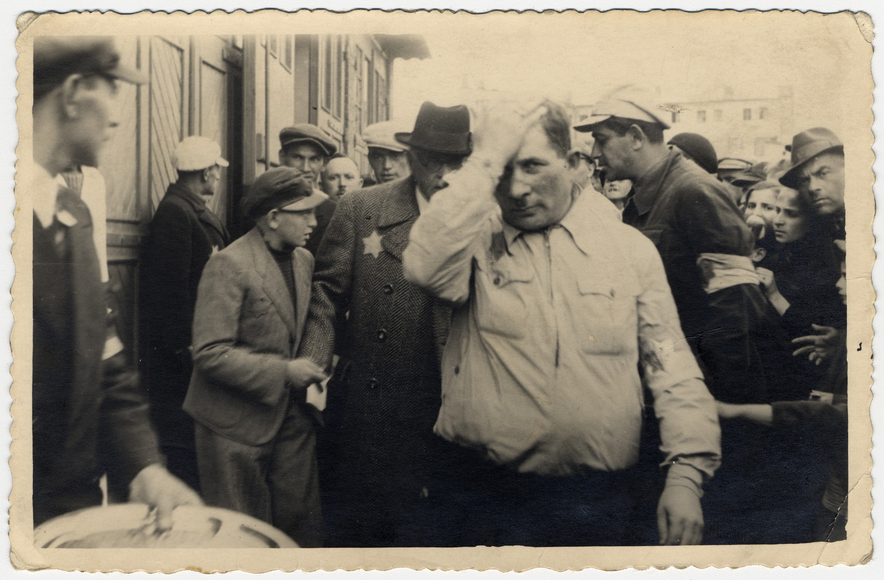 A young man hands a petition to Mordechai Chaim Rumkowski surrounded by other ghetto residents. 

The man in the foreground is Sergeant Kaufman.

The Yiddish inscription on the back reads: "Sergeant Kaufman from the Jewish guard, here protecting the Chairman M.Ch. Rumkowski, who allowed a chosen child to approach him with a petition.  August 17, 1941"