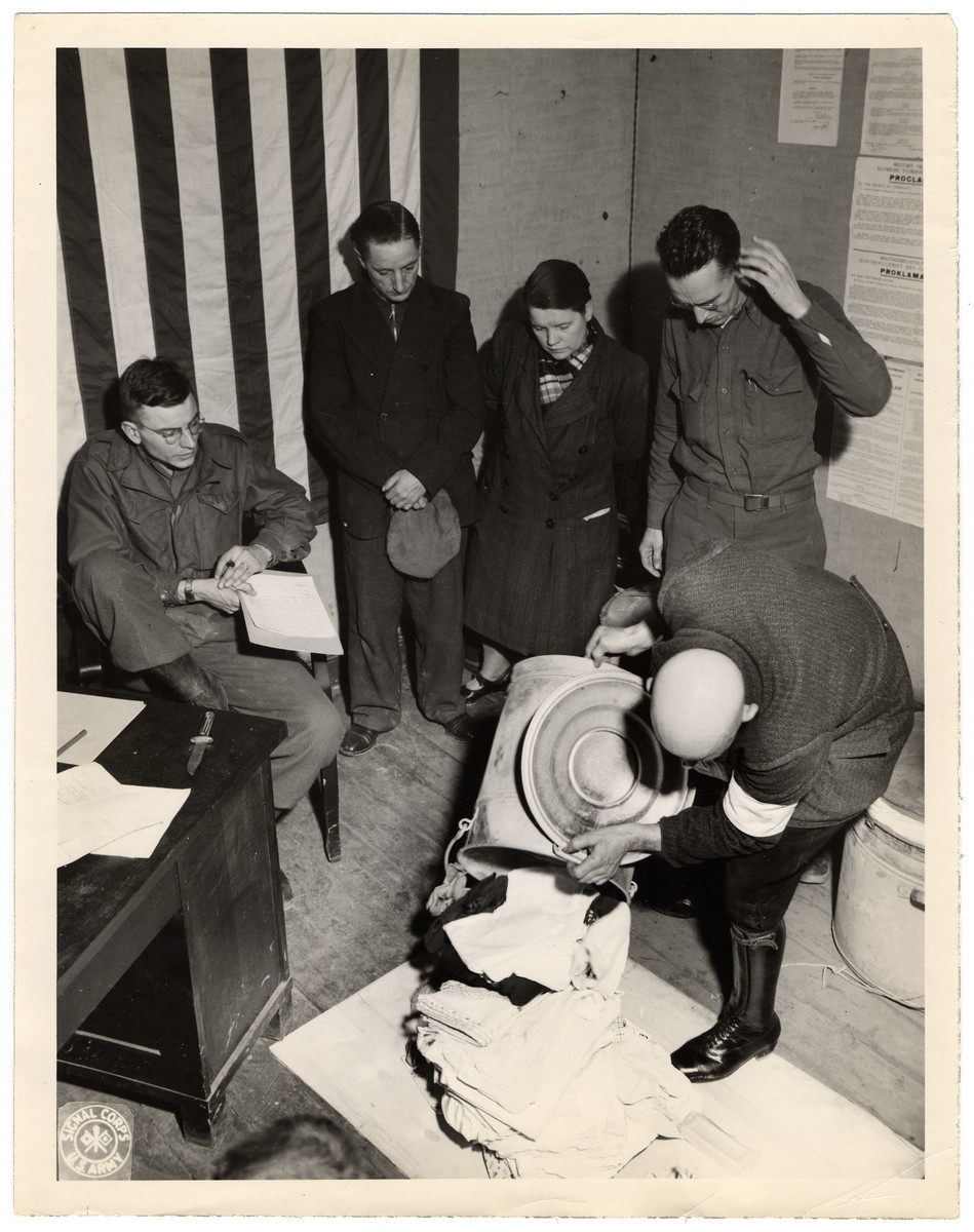 American soldiers oversee a court-martial of German civilians accused of looting.

Original Caption: "Court-martial trial of two German civilians accused of looting property from an abandoned and severely damaged house in Wuerselen. A civilian auxiliary policeman in exhibiting the property to the court. From left to right- the recorder, the two accused and the interpreter, then the civillian policeman."