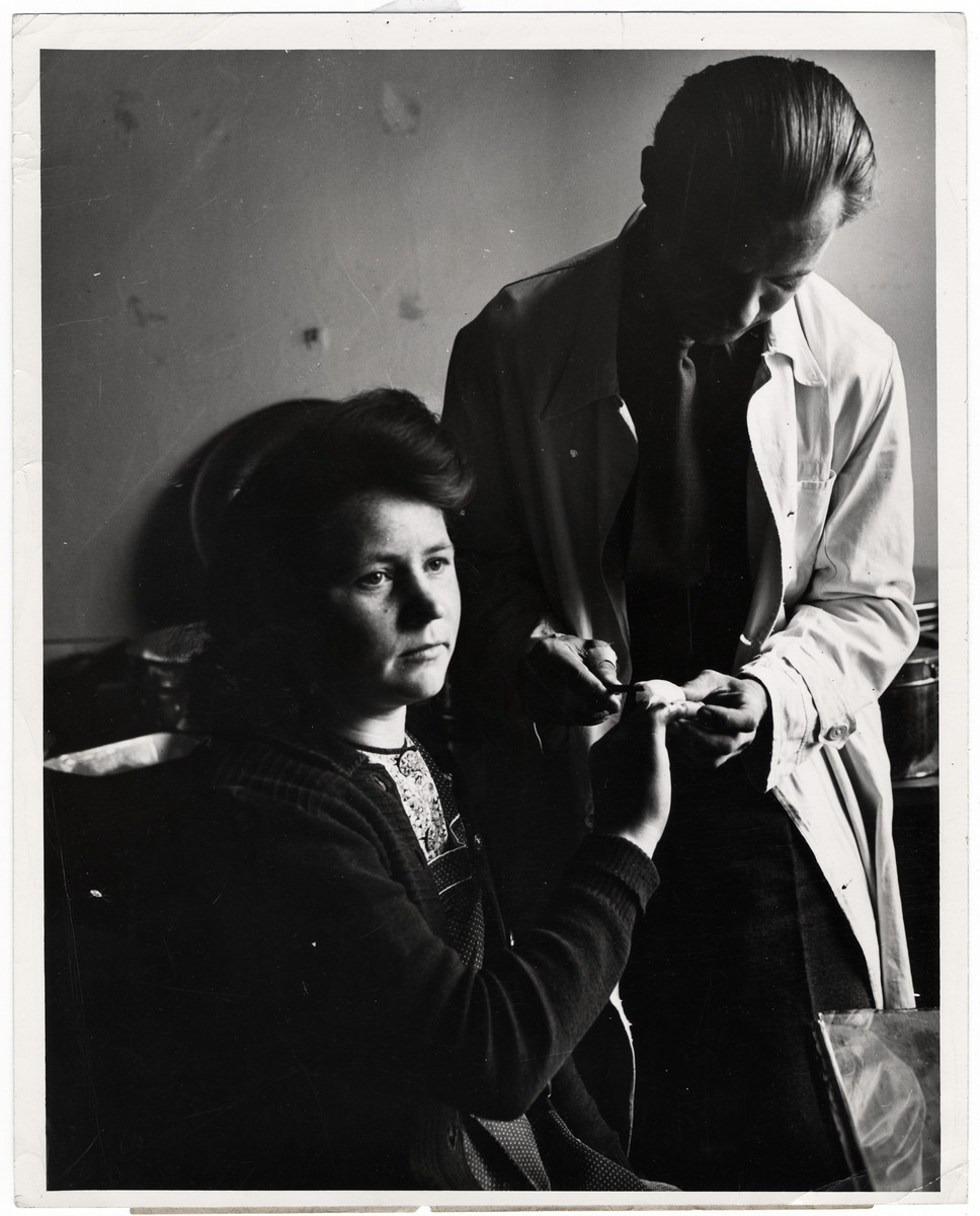 Orignial Caption: "Dr. Alphonse Kujawski of Possnau, Poland, applies a sterile dressing to a finger wound suffered by 21-year-old Stephania Kawozynska, who deported in 1940 with her family from Poland to slavery in Germany. 

When liberated, she was found with the badly infected finger, but amputation was avoided by careful treatment by the Allied doctors at the displaced persons center at Gnisnau Kasserne, a former German army post. All persons admitted into these camps are given thorough physical examinations and many are found to be suffering from a variety of ailments, including skin diseases. Liberated by the Allied armies in Germany, more that four million men, women and children, former slaves of the Nazi war machine, are free to return to their native lands as soon as repatriation machinery makes it possible. Meanwhile, these displaced persons are being gathered from the fields, factories and roadsides of Germany by military government authorities and placed in camps where they are registered, fed, and clothed, sheltered and given medical attention.

Persons of almost a dozen nationalities, living in separate camps wherever possible, are guided along democratic lines in the operation of their communities. U.S. Army officials set up the displaced persons centers, guard the exterior of the camps and turnover the guidance problems to international teams representing the Unted Nations Relief and Rehabilitaion Administration. Once all the displaced persons are repatriated, UNRRA's job in Germany is finished, as it has nothing to do with Germans or Military activities.

The Governments of Belgium, Holland, France and Luxembourg have already brought thousands of their nationals home. By May 17, 1945, nearly 800,000 liberated prisoners-of-war and political deportees were evacuated from German camps. The group included 603,940 French, 130,140 British and 62,989 Americans. All the Americans, 100,000 Frenchmen and 119,000 Britans were evactuated by air."