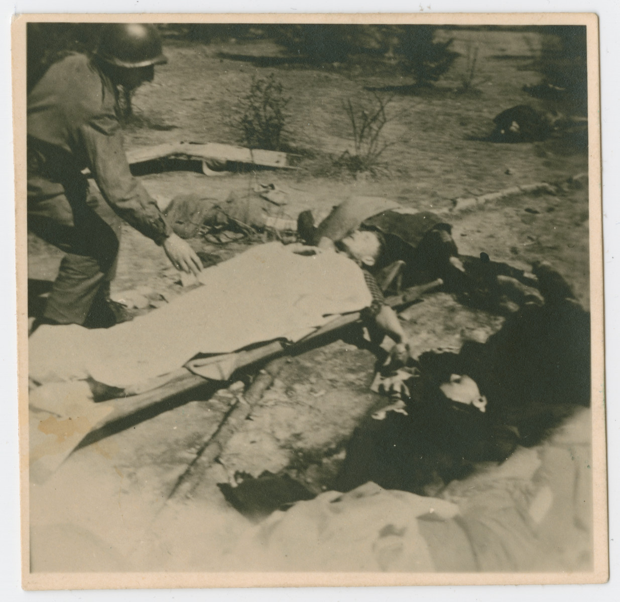 An American serviceman inspects the covered body of either a survivor or a victim lying on a stretcher on the grounds of the Ohrdruf concentration camp.