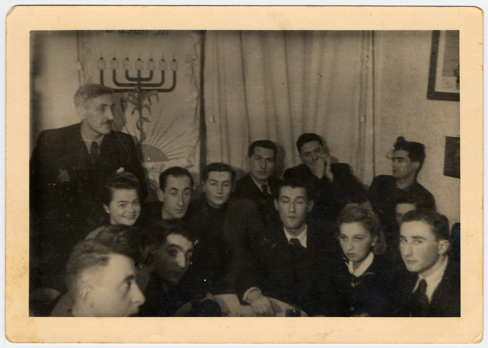 Members of a Zionist group in the Lodz ghetto at a Hanukkah gathering.

Among those pictured is Yehoshua Klugman (standing, left).  Seating in the first row are Ester Grynsztajn (first from left) and Aron Jakubson (second from left in the second row).  Seated in the first row on the right are Wiktor Sztajn, Mirka Sztajn and Libicki.  Pictured seated in the second row on the right are Chaim Rozenowicz and next to him, against the wall, is Arie Princ (later Ben Menachem).  The banner in the back reads: "1943--Festival of the Maccabees."  Latin letters on the branches of the menorah spell "KASPIJK," possibly the name of the group or chapter of the organization.