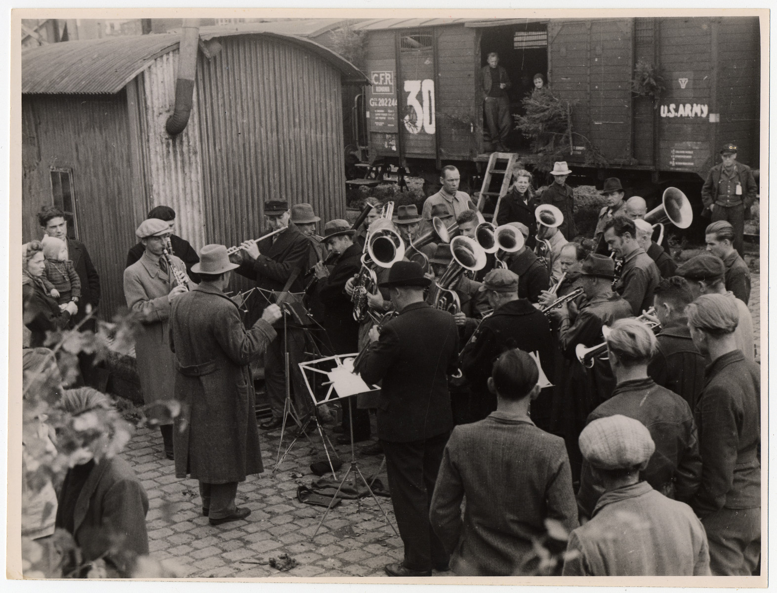 An orchestra performs at a train station as a send-off to a group of displaced persons leaving on a U.S. army train.