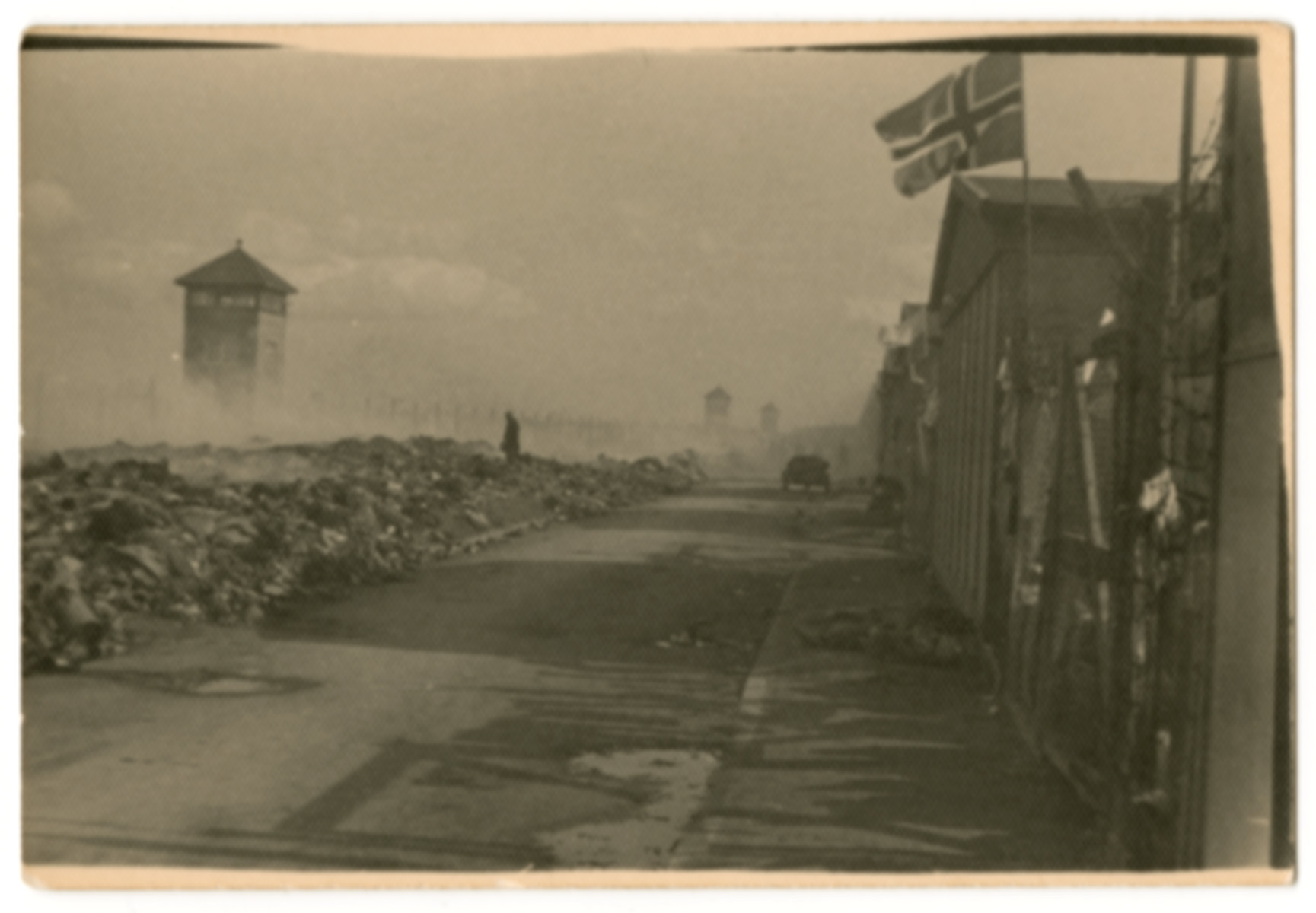 A Norwegian flag flys from a barrack in Dachau facing a mound of rubble and a watch tower.